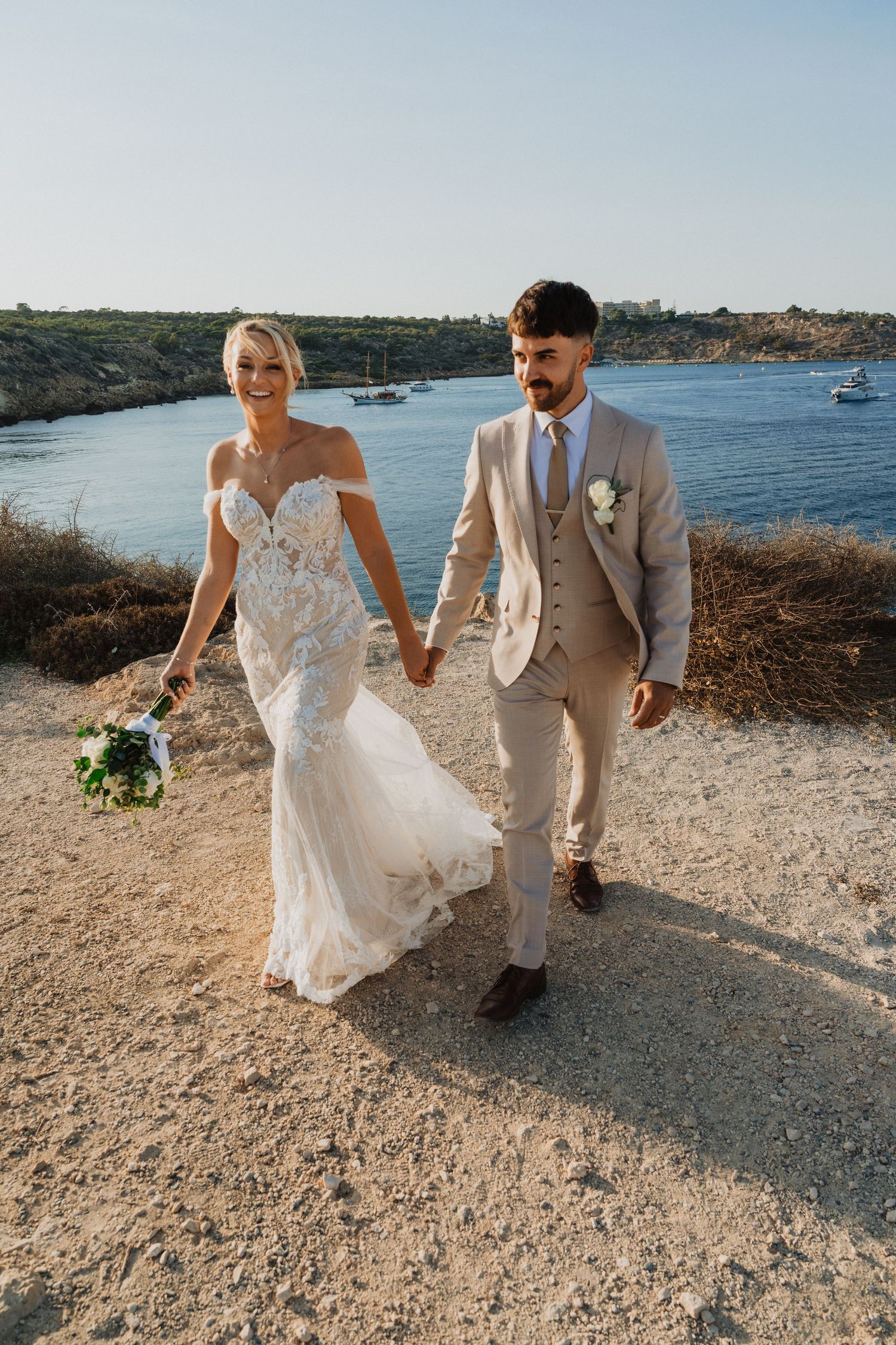 Bride in lace gown and groom in tan suit hold hands while walking along coastal path.