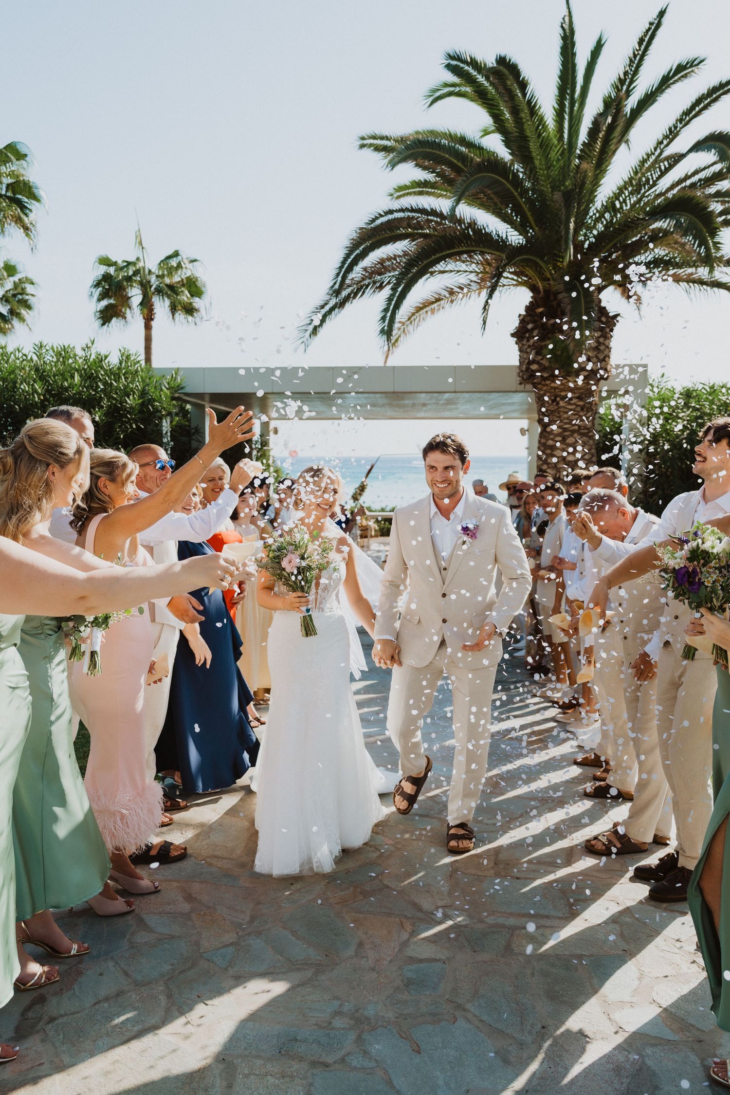 Wedding guests create a festive atmosphere throwing confetti along a palm-lined pathway.