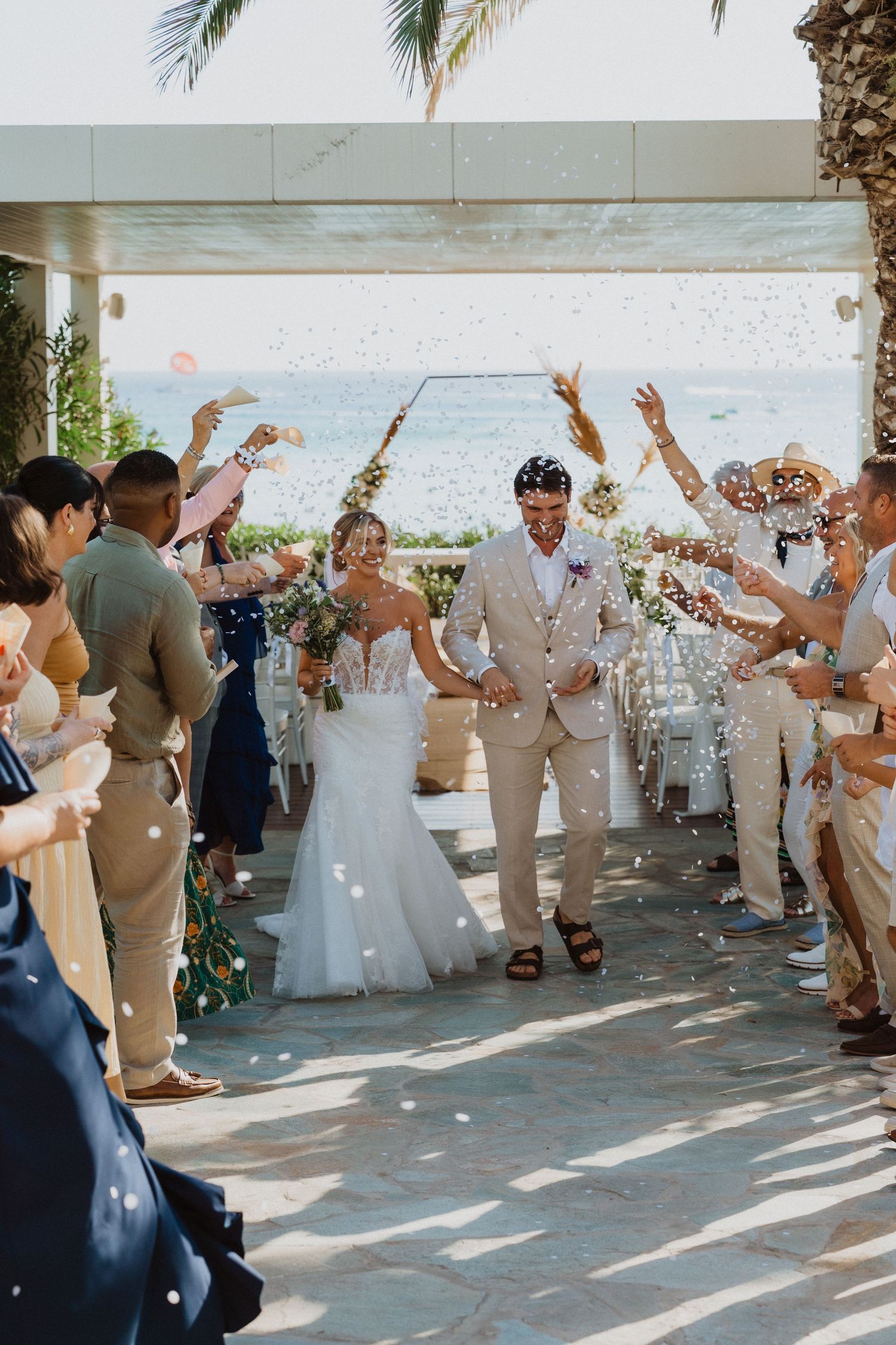 Wedding guests throw confetti as newlyweds walk through an outdoor venue with palm trees.