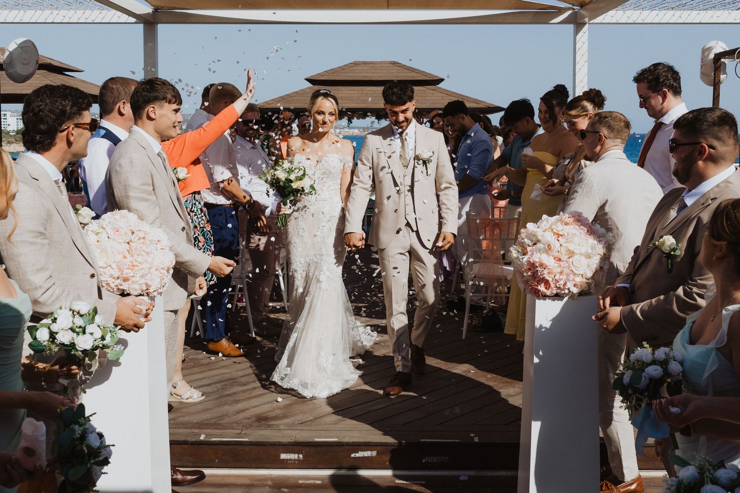 Wedding party and guests gather for celebration under white canopy by the ocean.