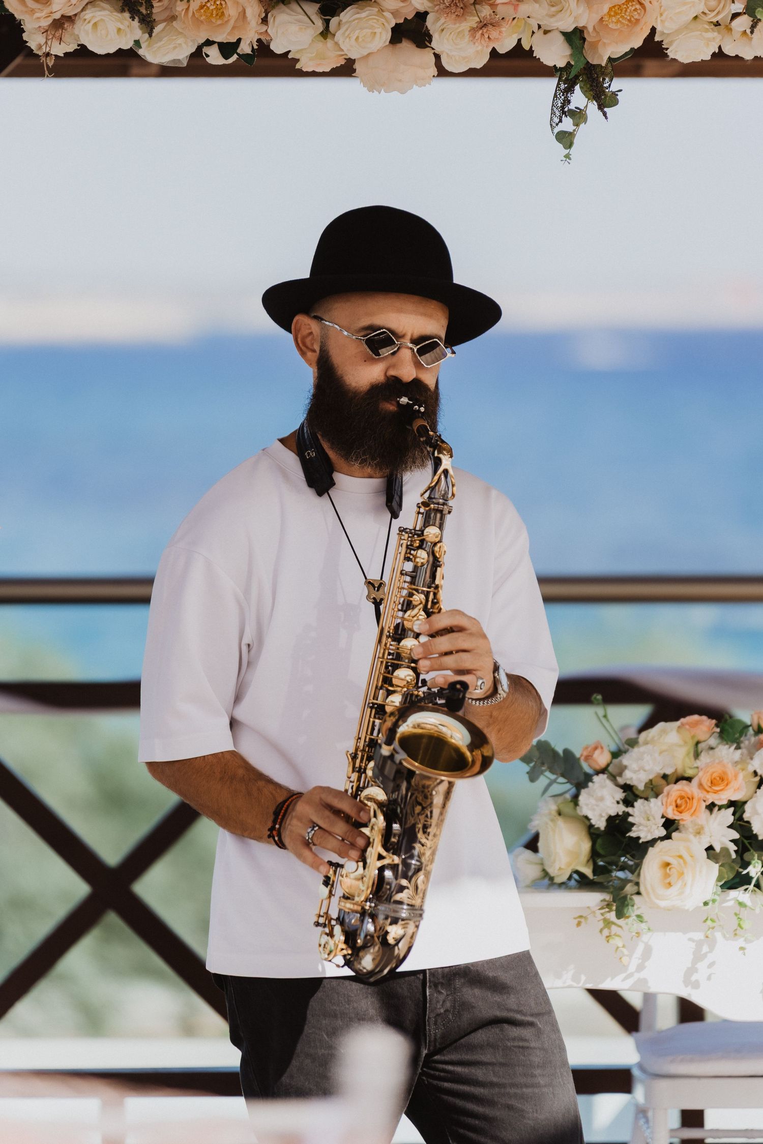 Close-up of saxophone performer in white shirt and black hat with floral decorations and ocean background.