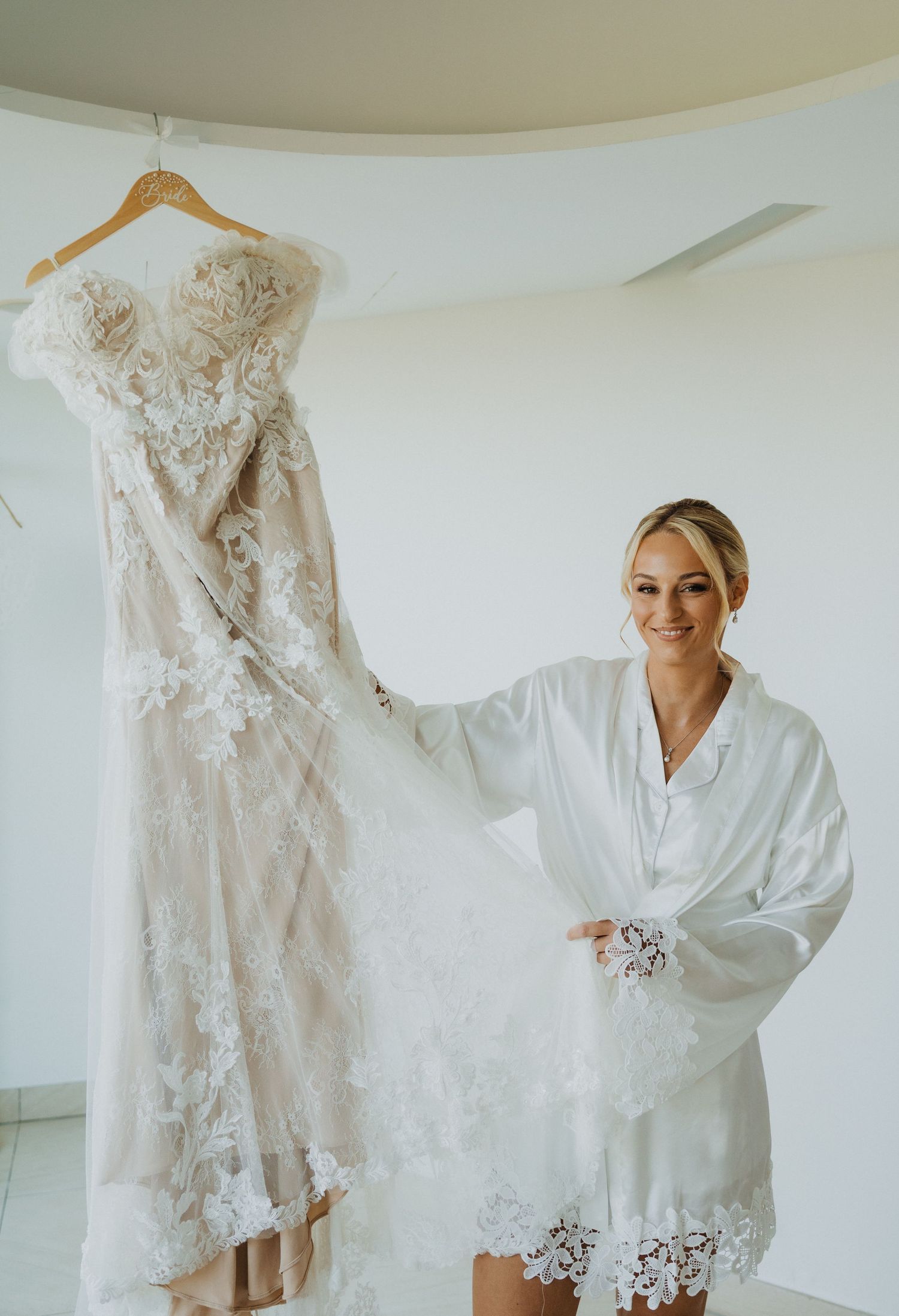 A white lace wedding dress hangs on a wooden hanger while someone in a white robe stands nearby.