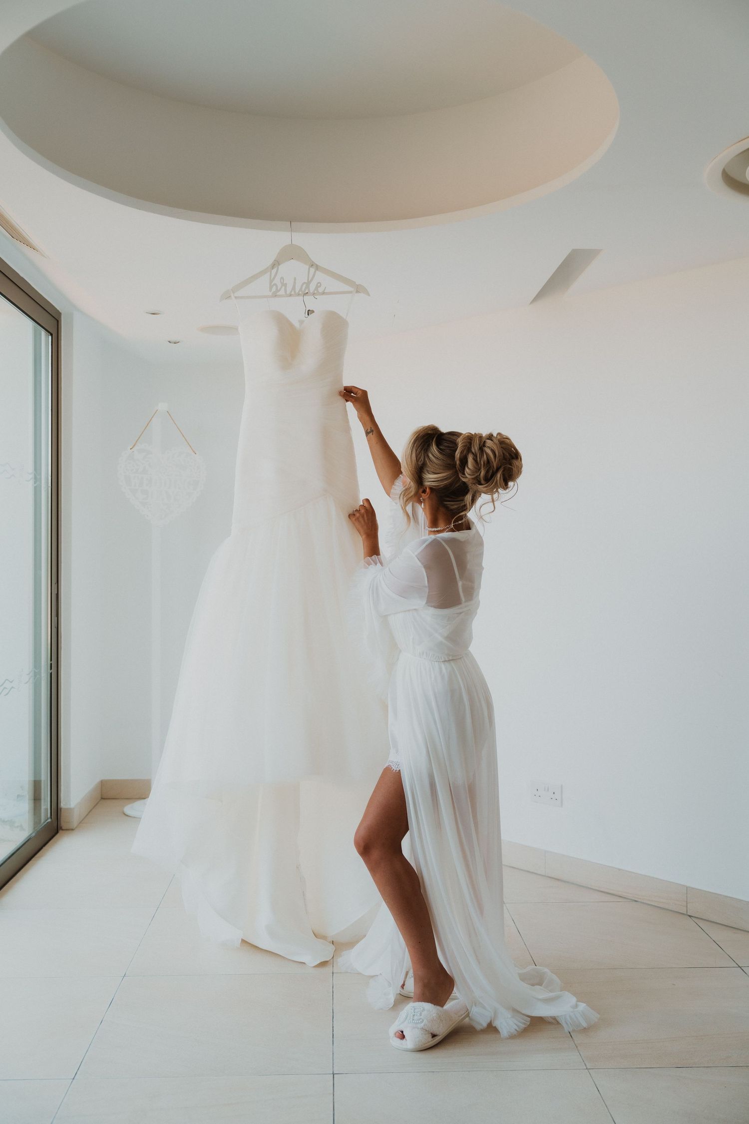 Bride in white robe reaching up to touch her wedding gown hanging in a bright modern bridal suite.