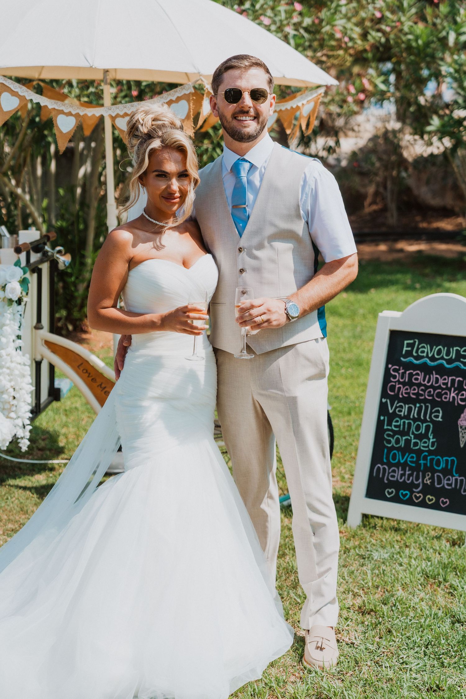 Bride in white mermaid gown and groom in tan suit pose together at outdoor wedding reception.