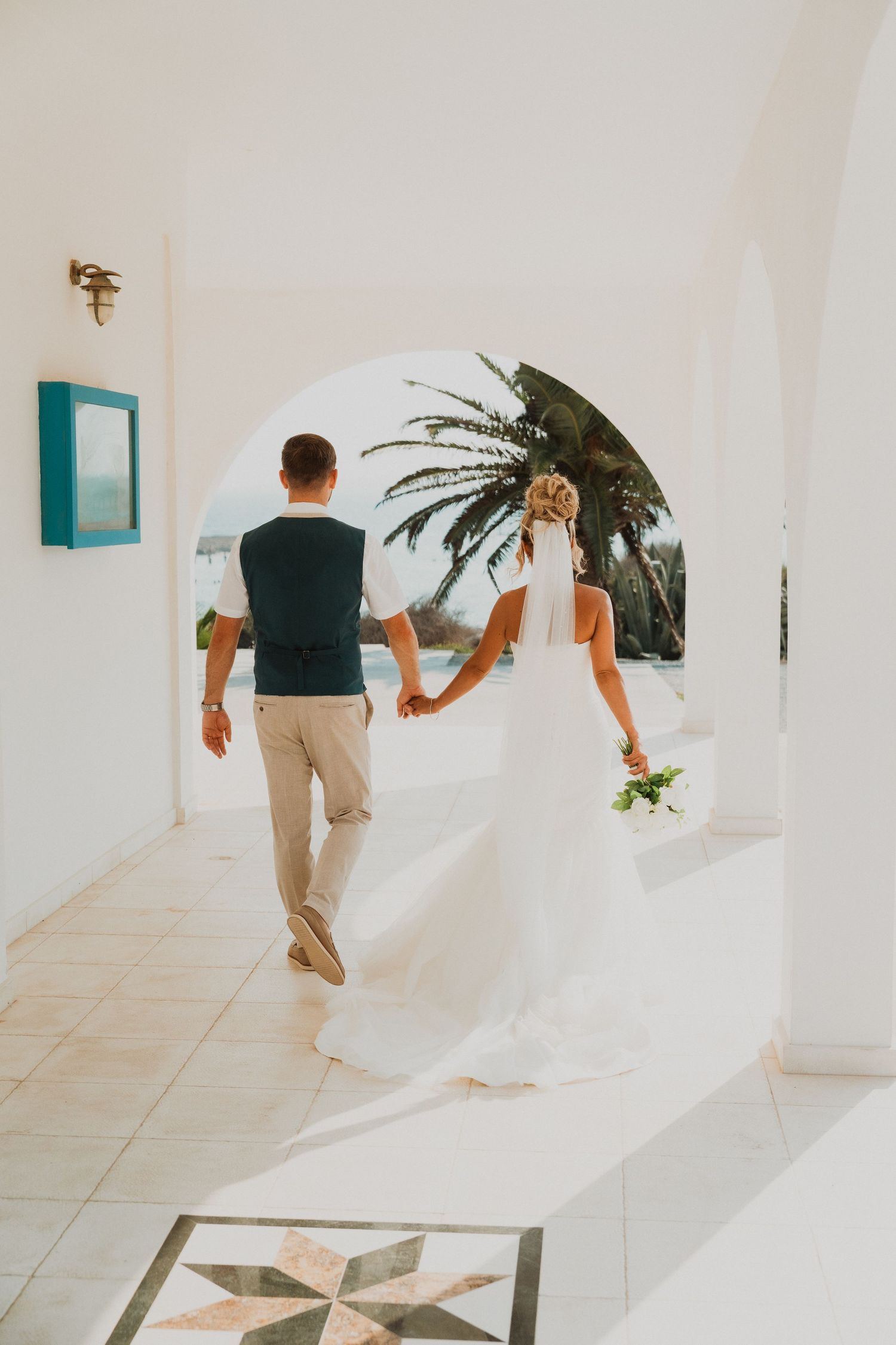 Bride and groom holding hands walking through white arch toward palm trees at a Mediterranean wedding venue.