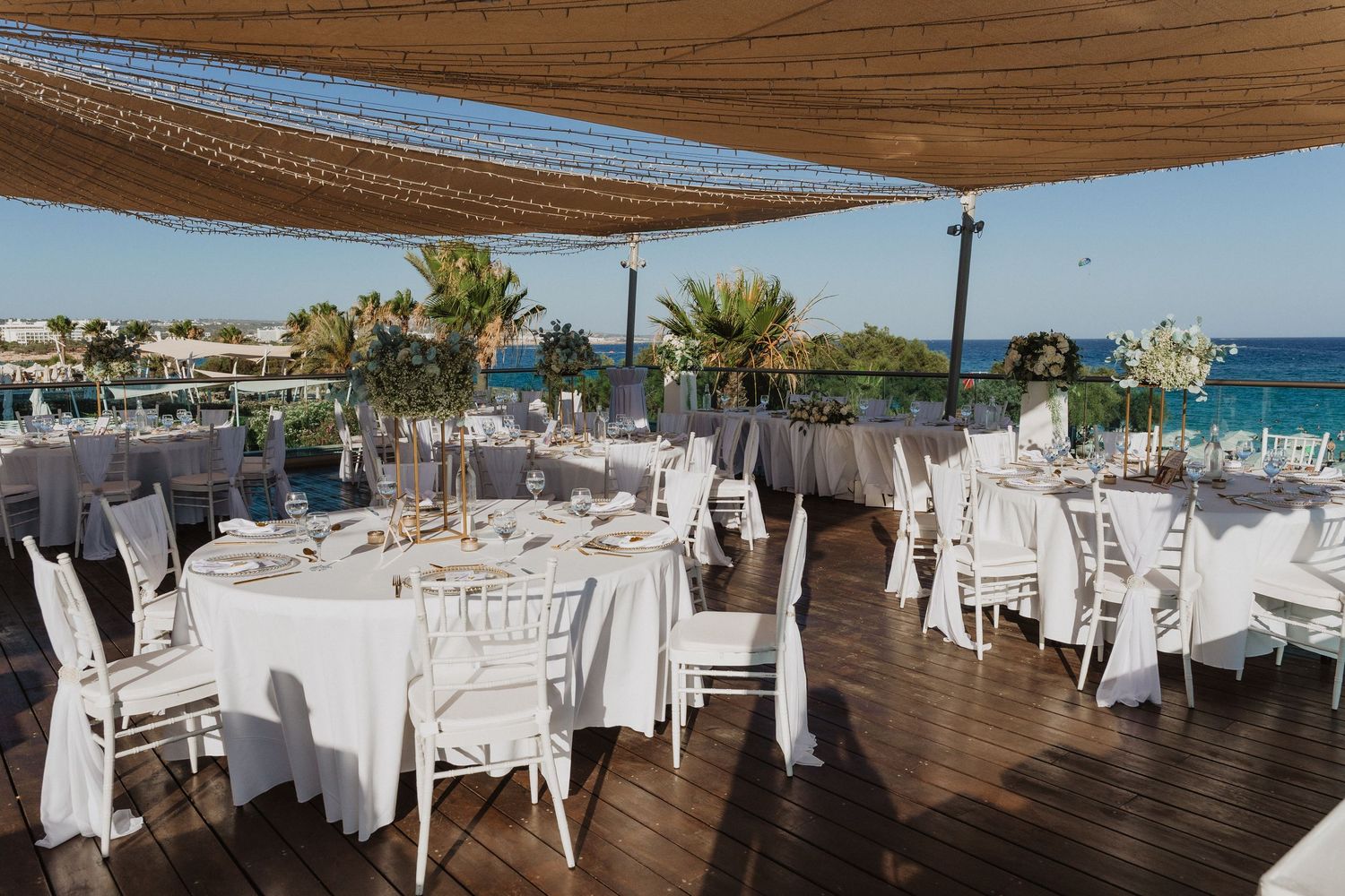 Elegant outdoor wedding reception setup with white tables and chairs on a wooden deck overlooking the ocean.