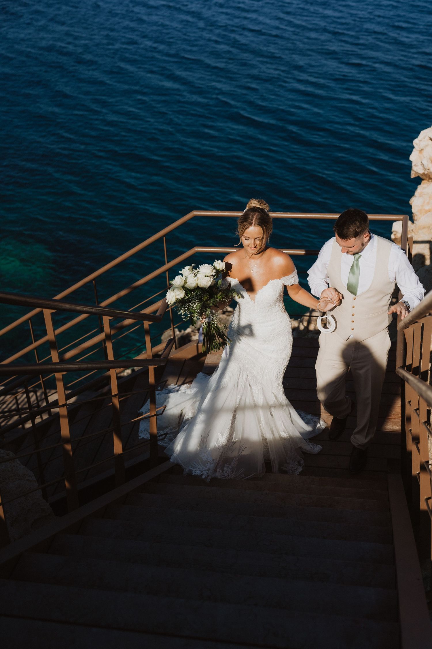 A couple in wedding attire descending outdoor stairs near deep blue water at sunset.
