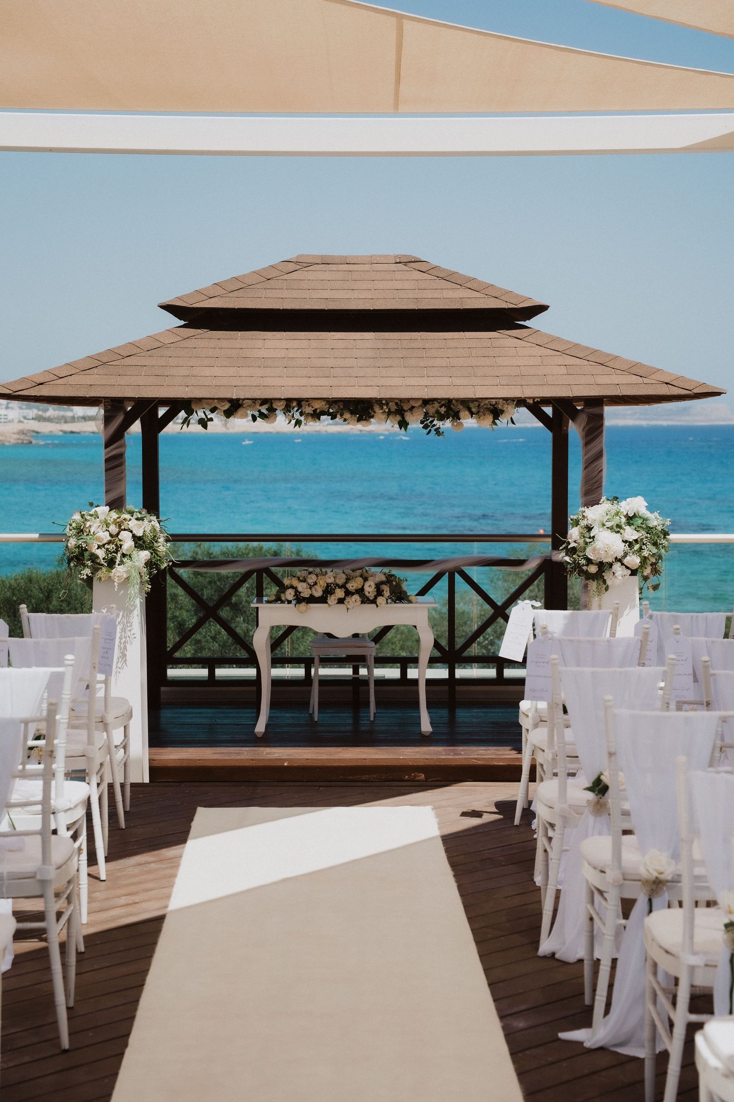 A beachside wedding ceremony setup with white chairs and wooden gazebos overlooking turquoise waters and blue skies.