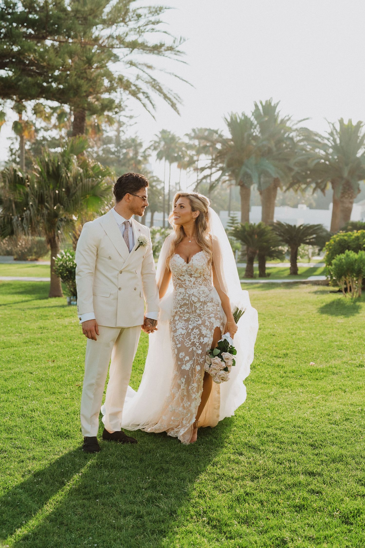 A couple in wedding attire walking together on a lush green lawn with palm trees in the background during sunset.