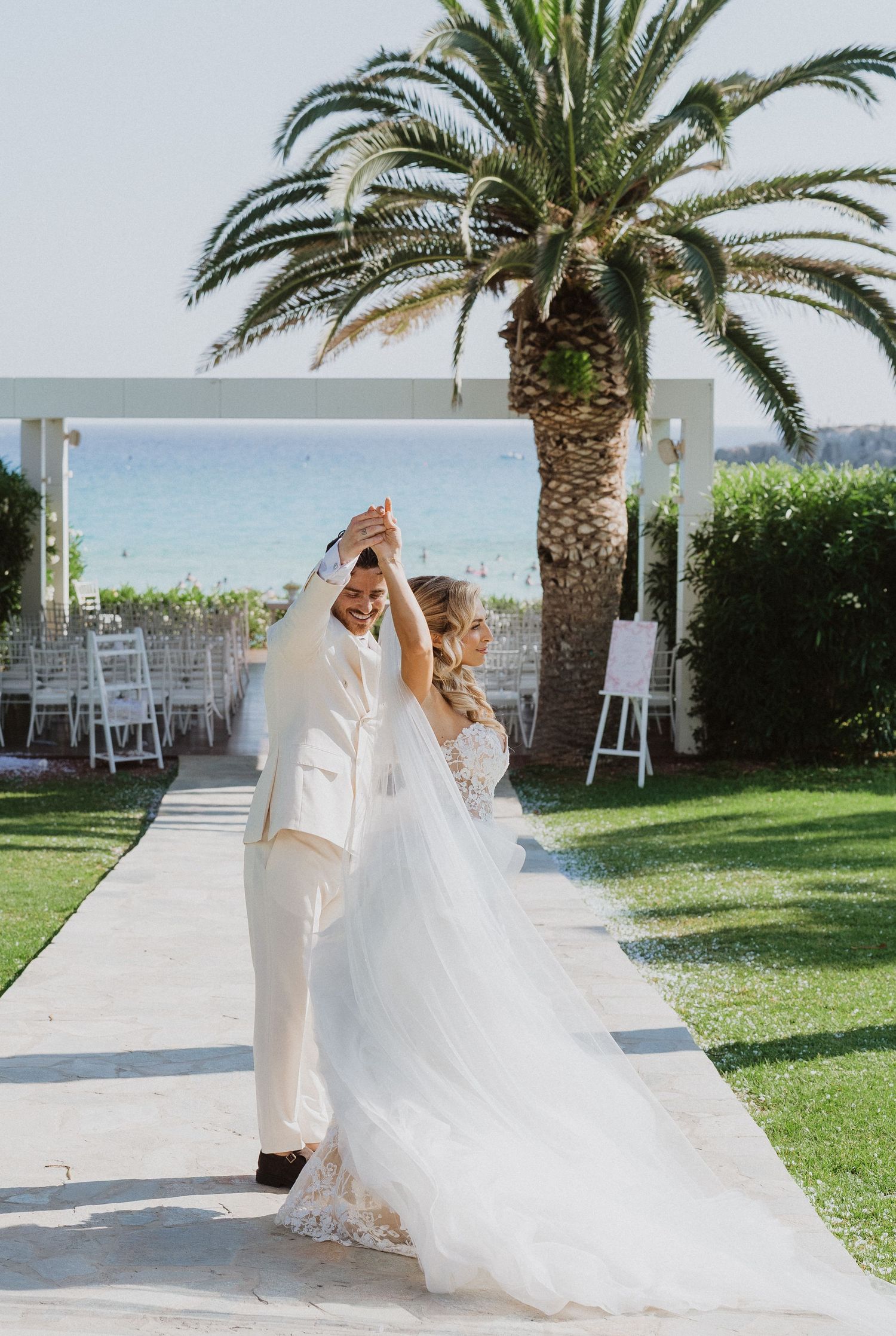 A wedding couple dancing on an outdoor pathway by the ocean with a palm tree and white chairs in the background.