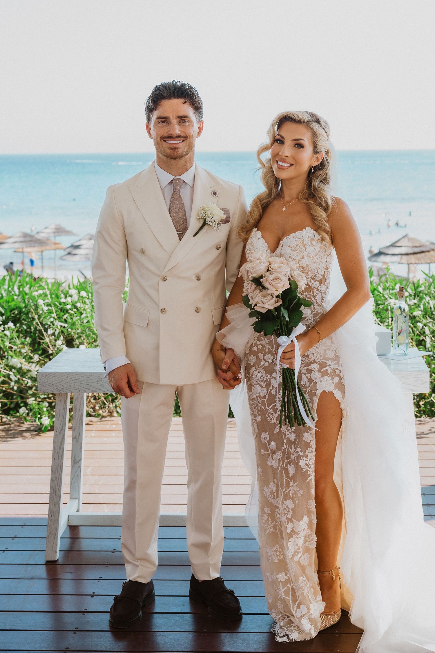 A couple in beige and lace attire poses together for beach wedding photos on a wooden deck overlooking the ocean.