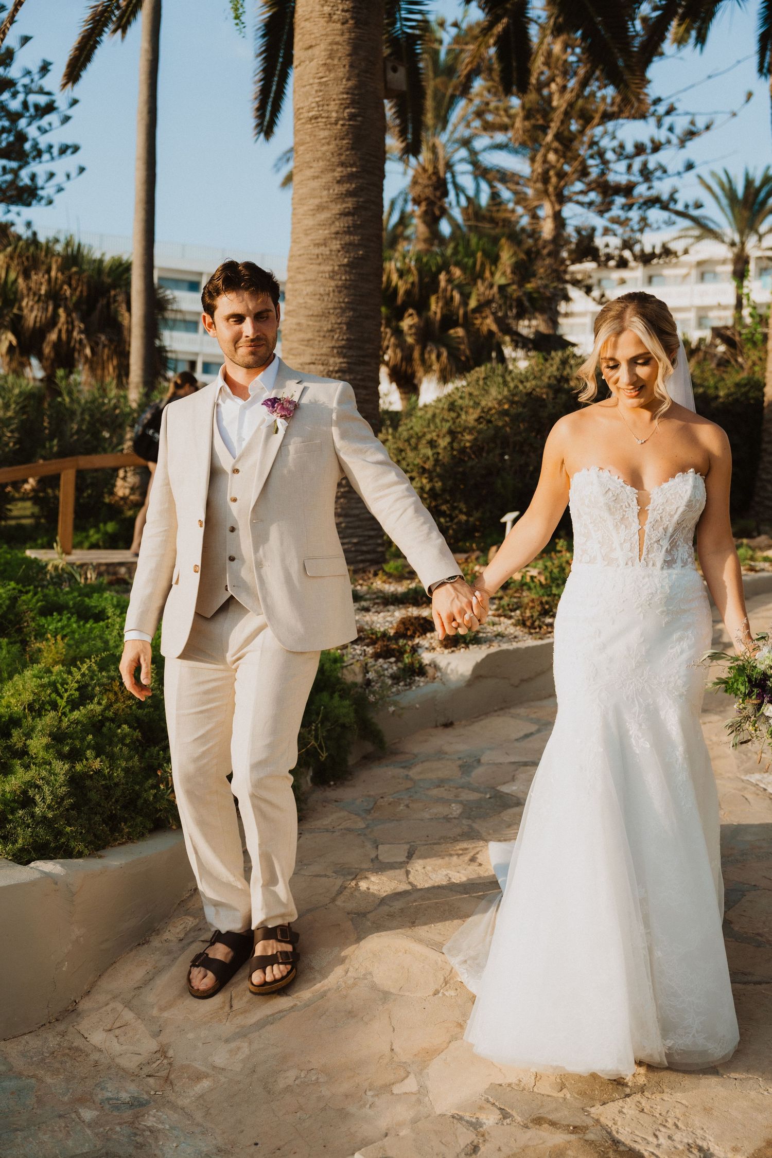 Elegant wedding couple strolls hand in hand through a palm-lined coastal garden.