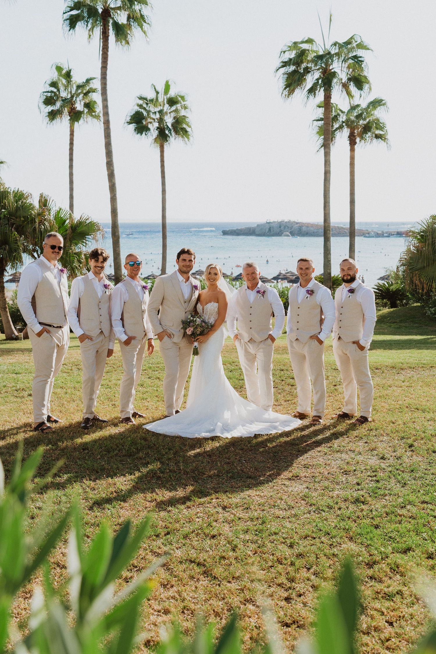 Wedding party in all-white attire poses under palm trees by the ocean.