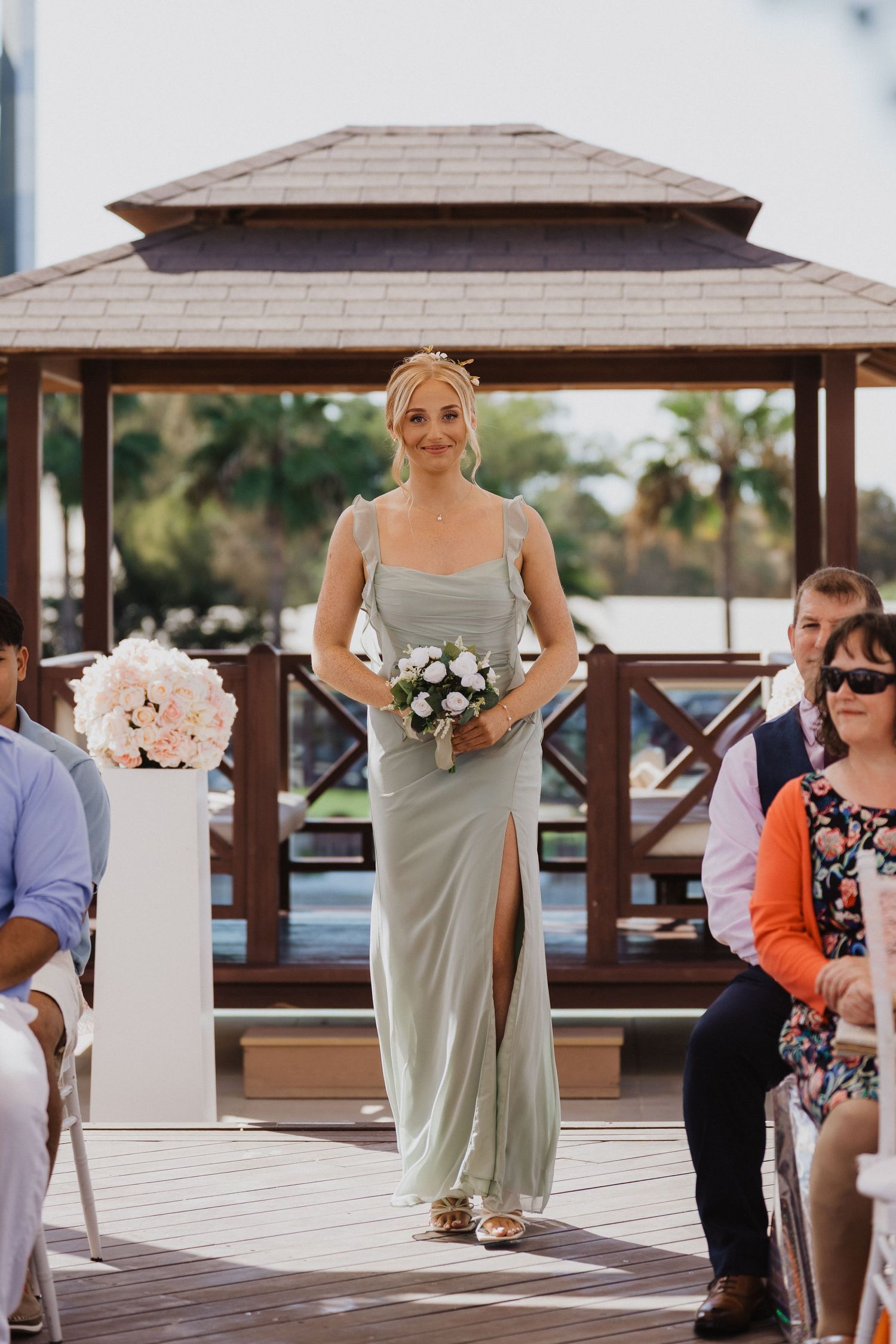 Wedding party members in sage green dresses walk down a wooden deck aisle carrying bouquets.