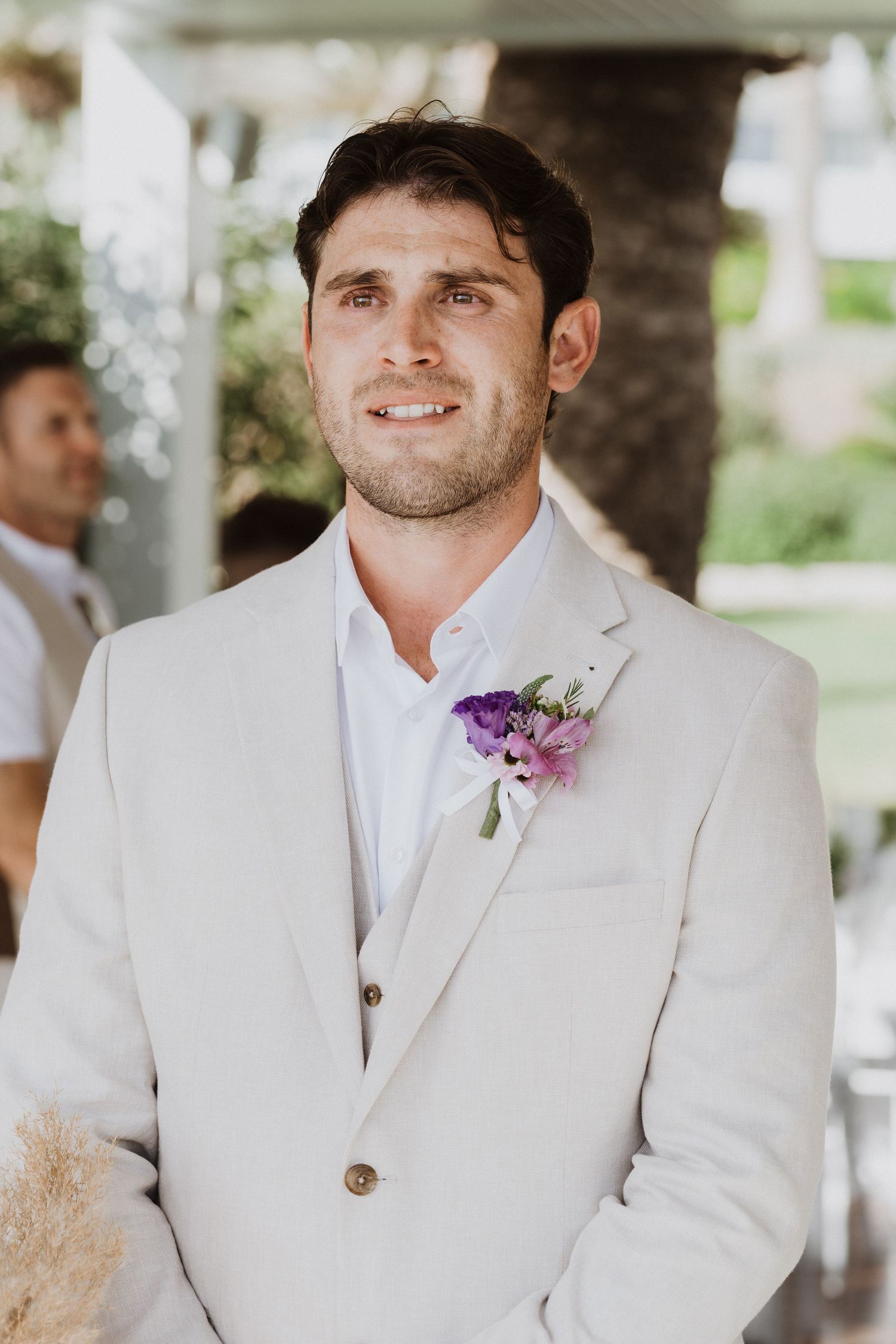 Groom wearing white suit with purple boutonniere at outdoor ceremony.