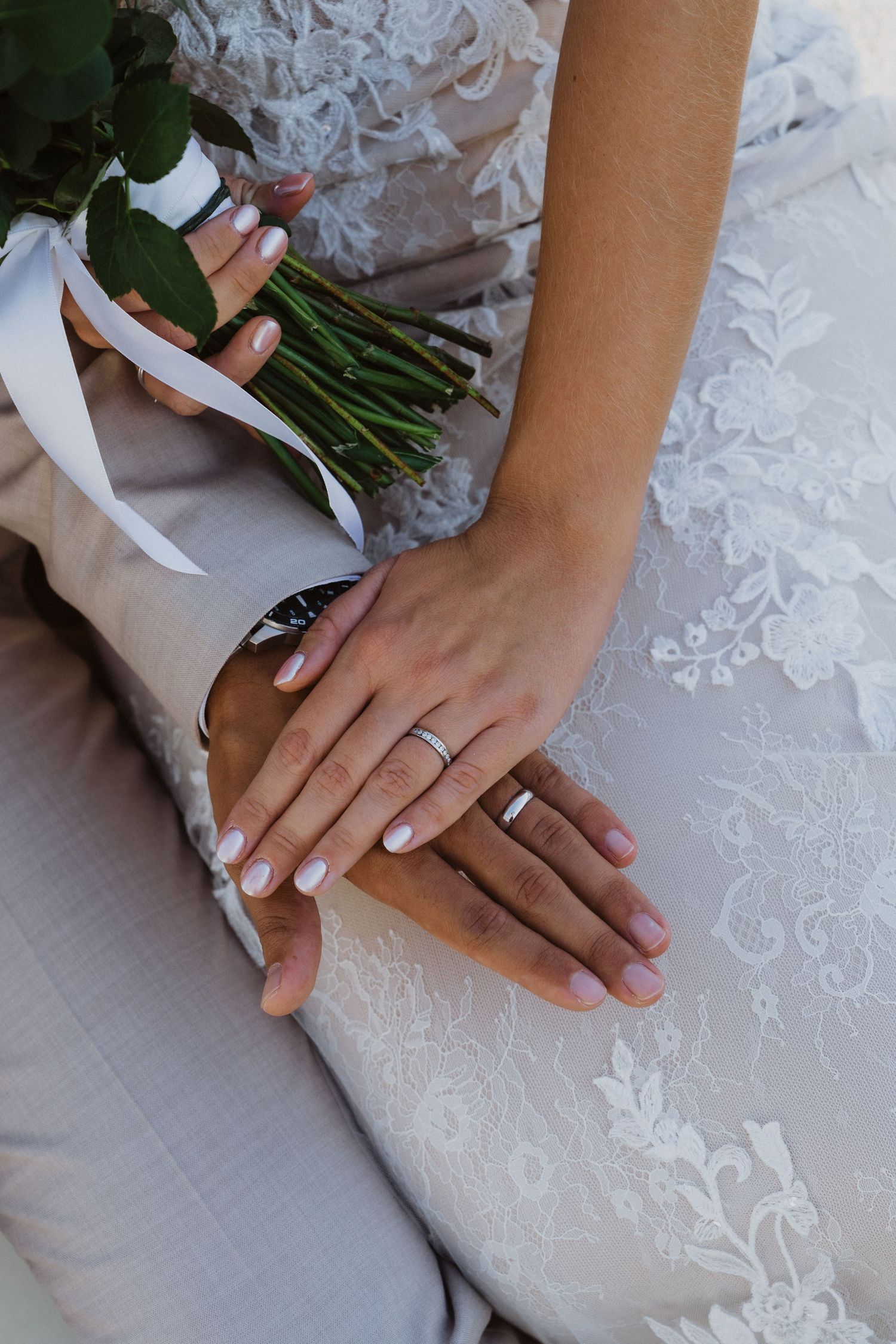 Close-up of wedding rings and manicured hands on white lace dress fabric.