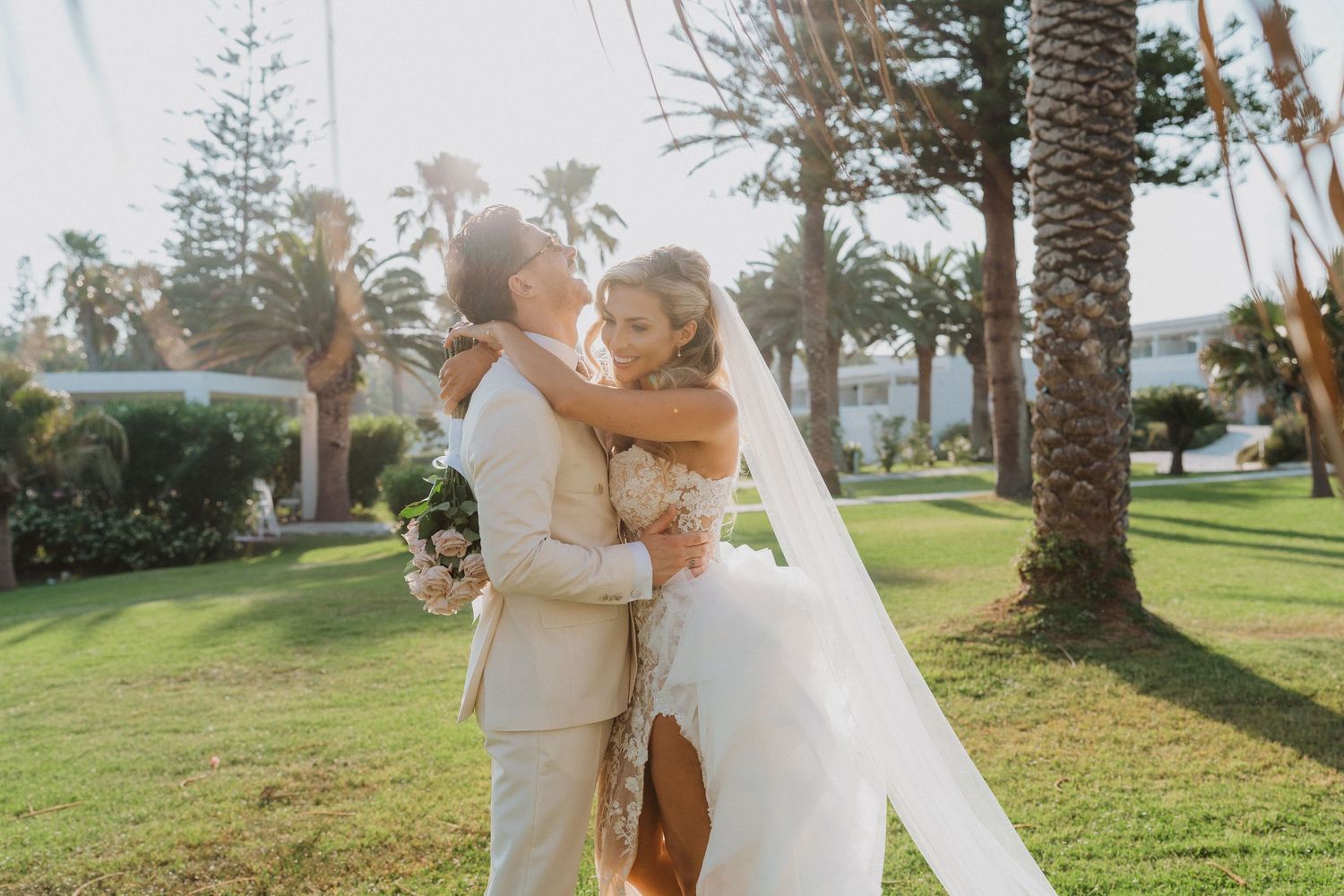 A romantic wedding moment captured in a lush tropical garden setting with palm trees at sunset.