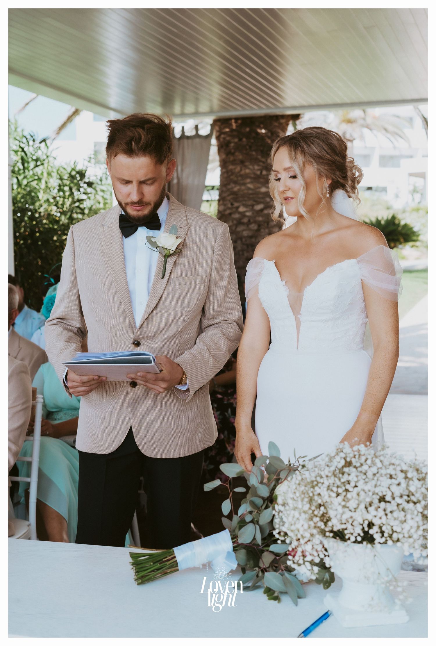 A couple reads from a document during their garden wedding ceremony with white floral decorations.