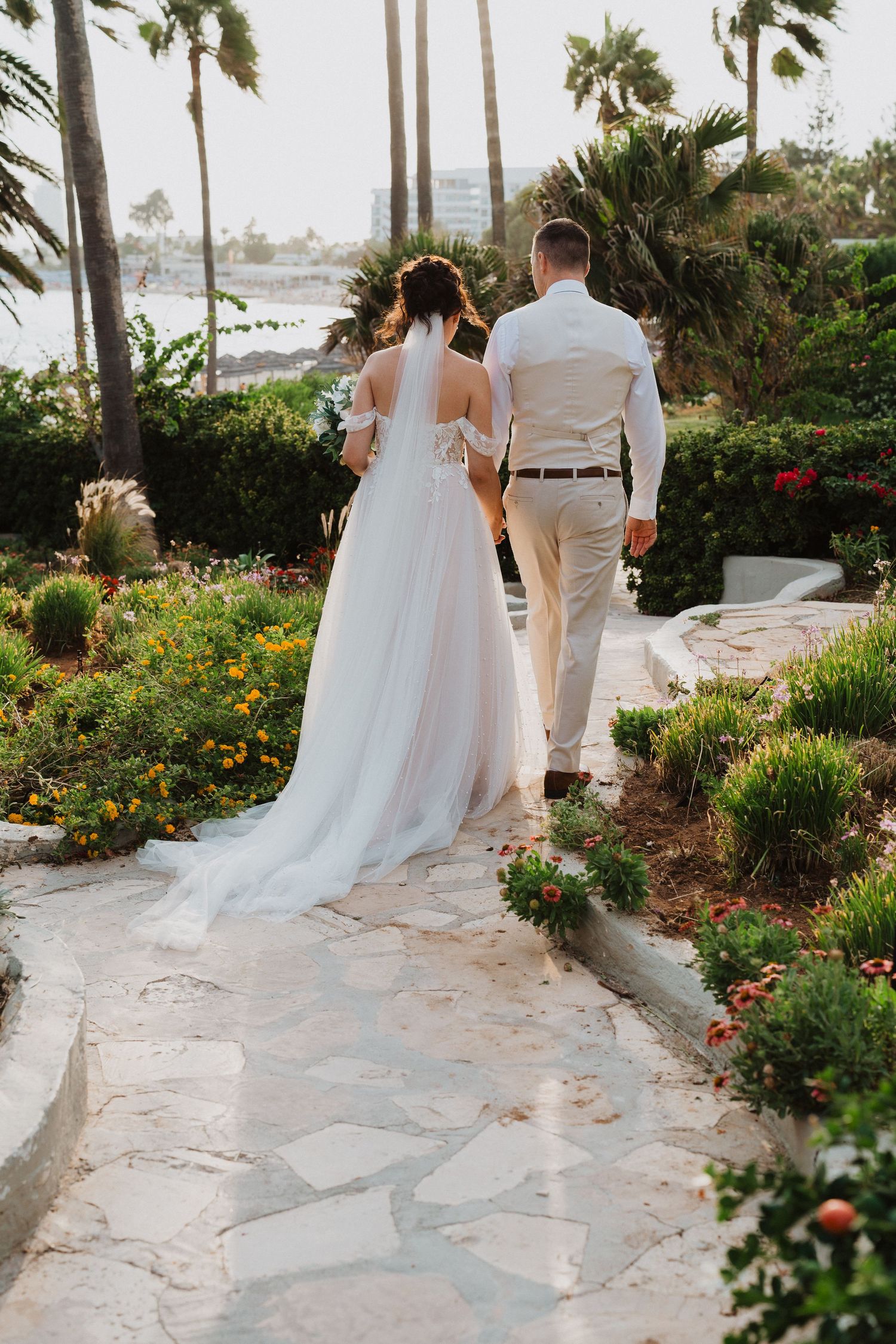 Newlyweds walk along a scenic stone path surrounded by tropical gardens and palm trees at a beachside venue.