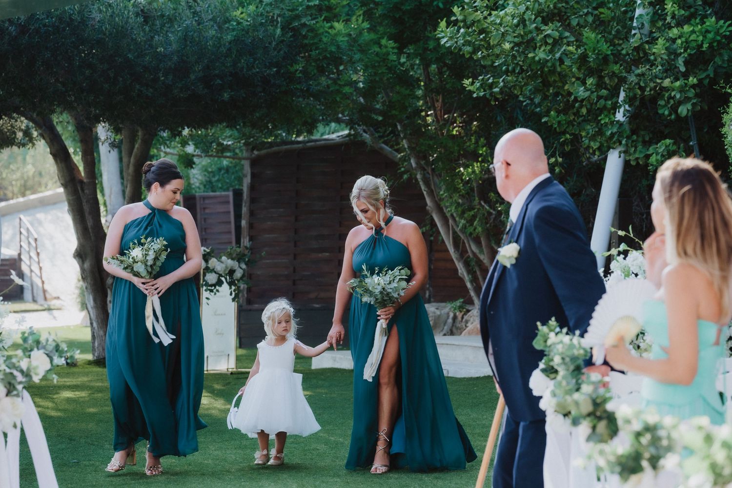 Bridal party stands outdoors on a lawn during wedding ceremony surrounded by greenery and floral decorations.