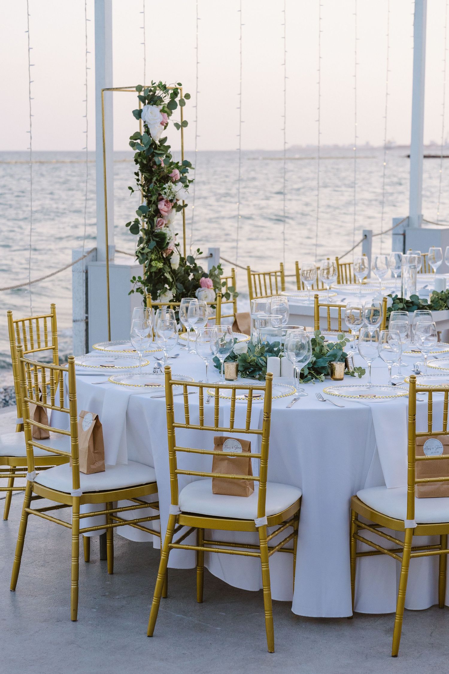 Elegant round dining tables with gold chairs and greenery centerpieces overlooking the sea.