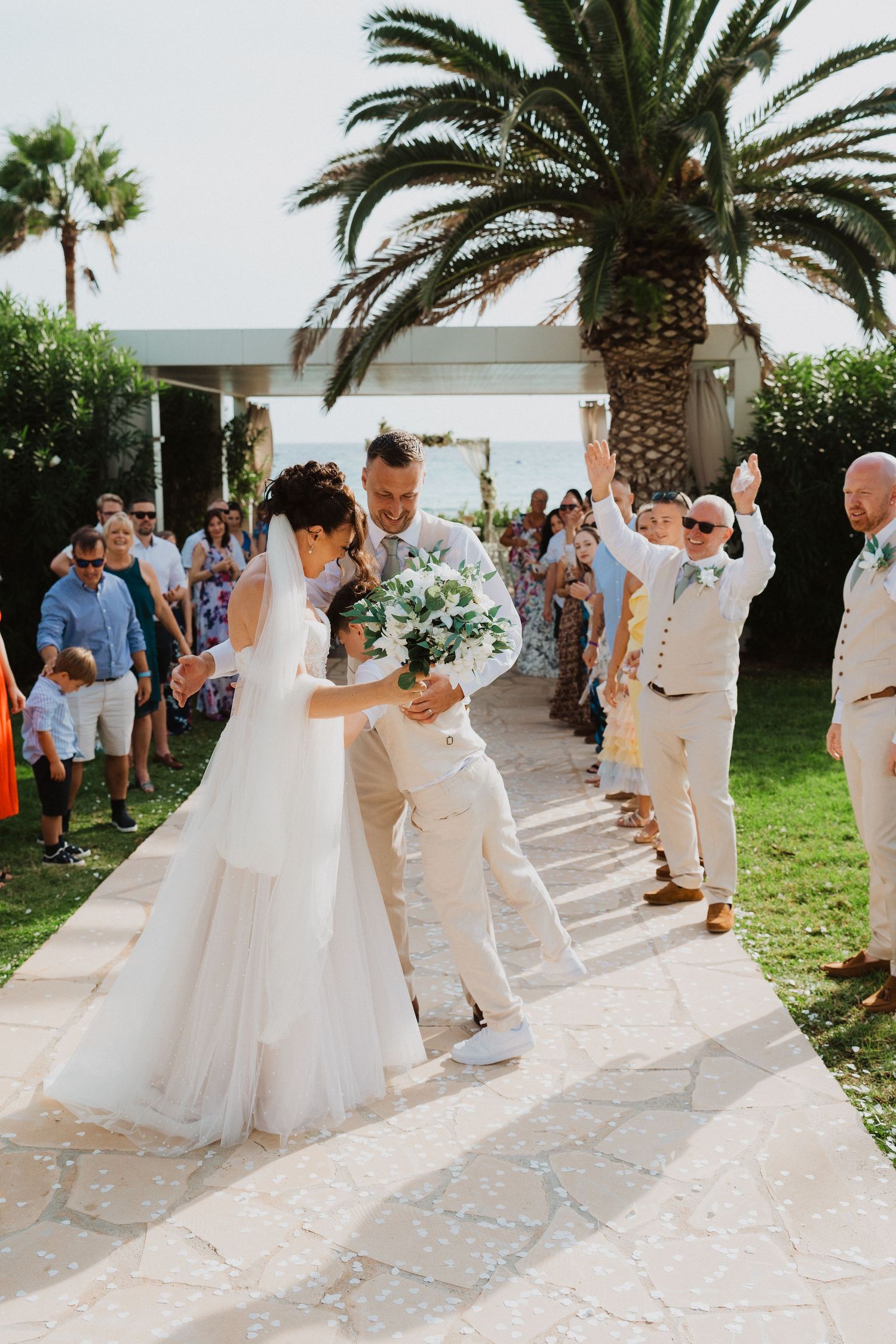A joyful wedding celebration takes place outdoors with a palm tree backdrop as guests gather on a stone pathway.