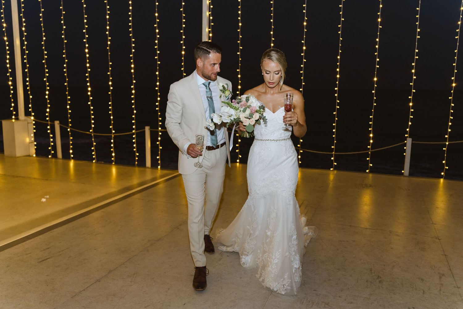 Newlyweds walk together holding drinks under romantic string light decorations.