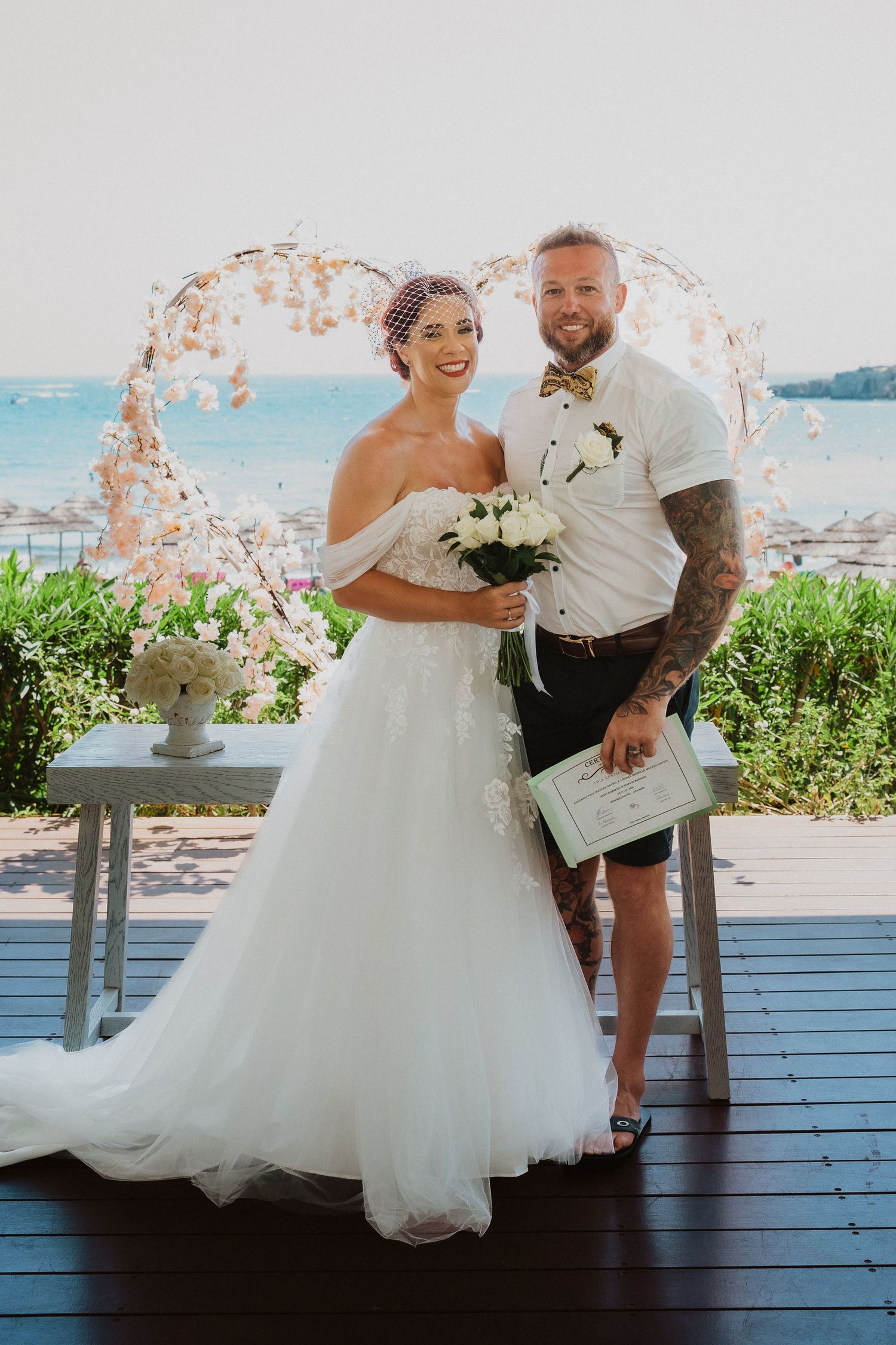 Newlyweds pose under a delicate floral arch against an ocean backdrop on their wedding day.