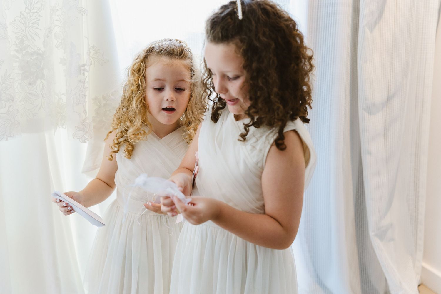Two people in white dresses share a tender moment while getting ready for a formal occasion.