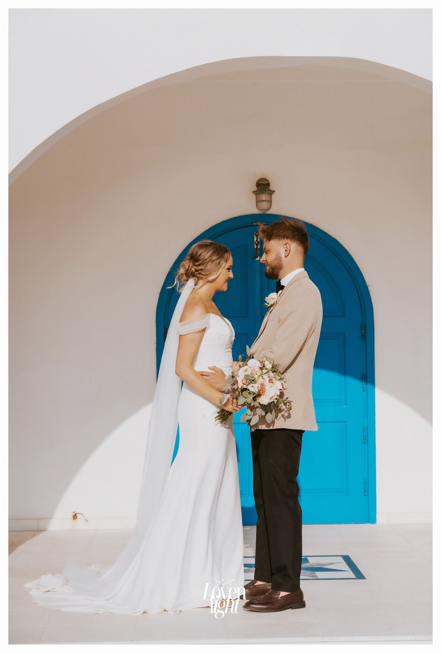 A romantic wedding portrait of a couple posed under a white archway with a bright blue door in the background.