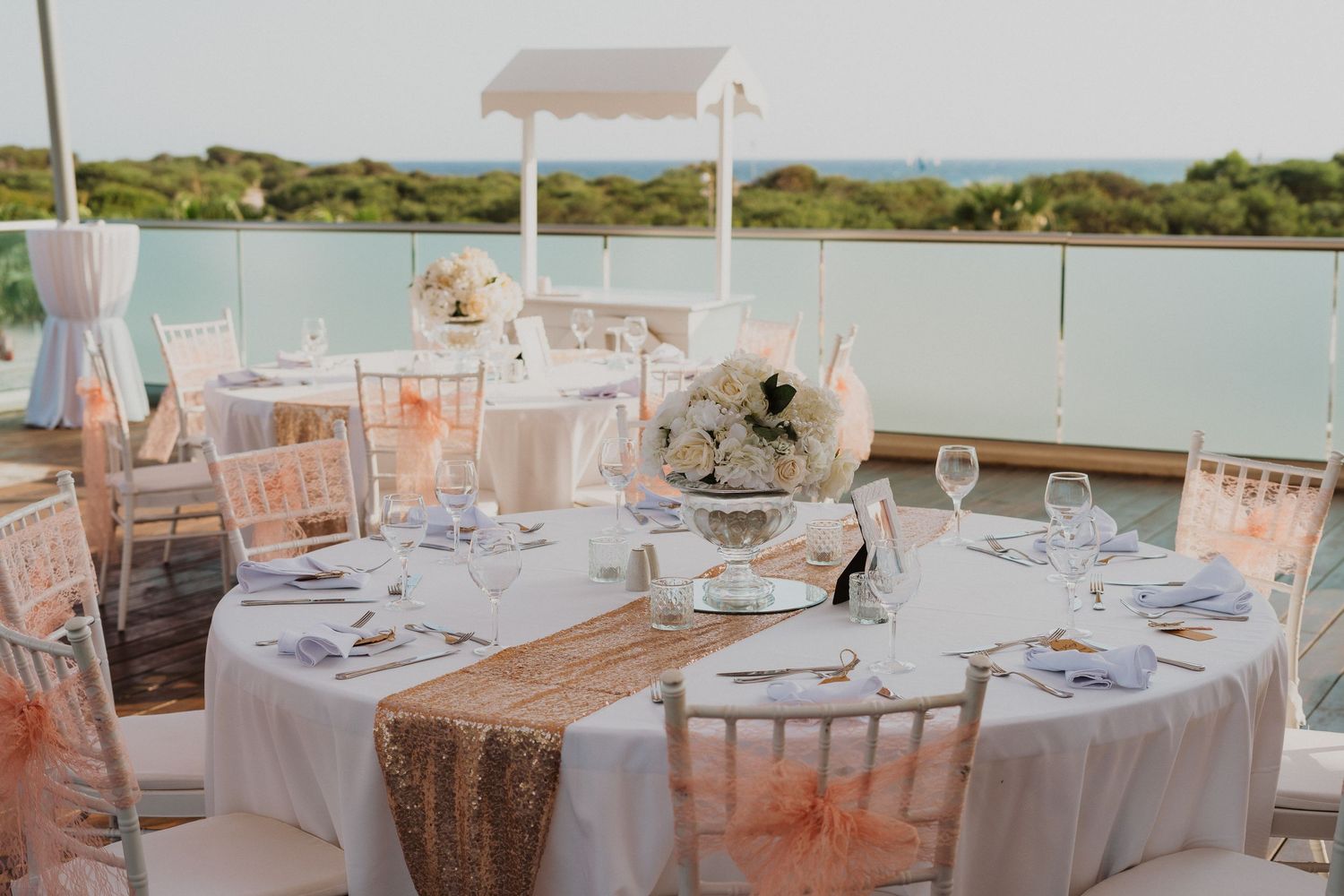 Romantic table setting with rose gold sequin runner, white linens and floral centerpiece at outdoor wedding venue.