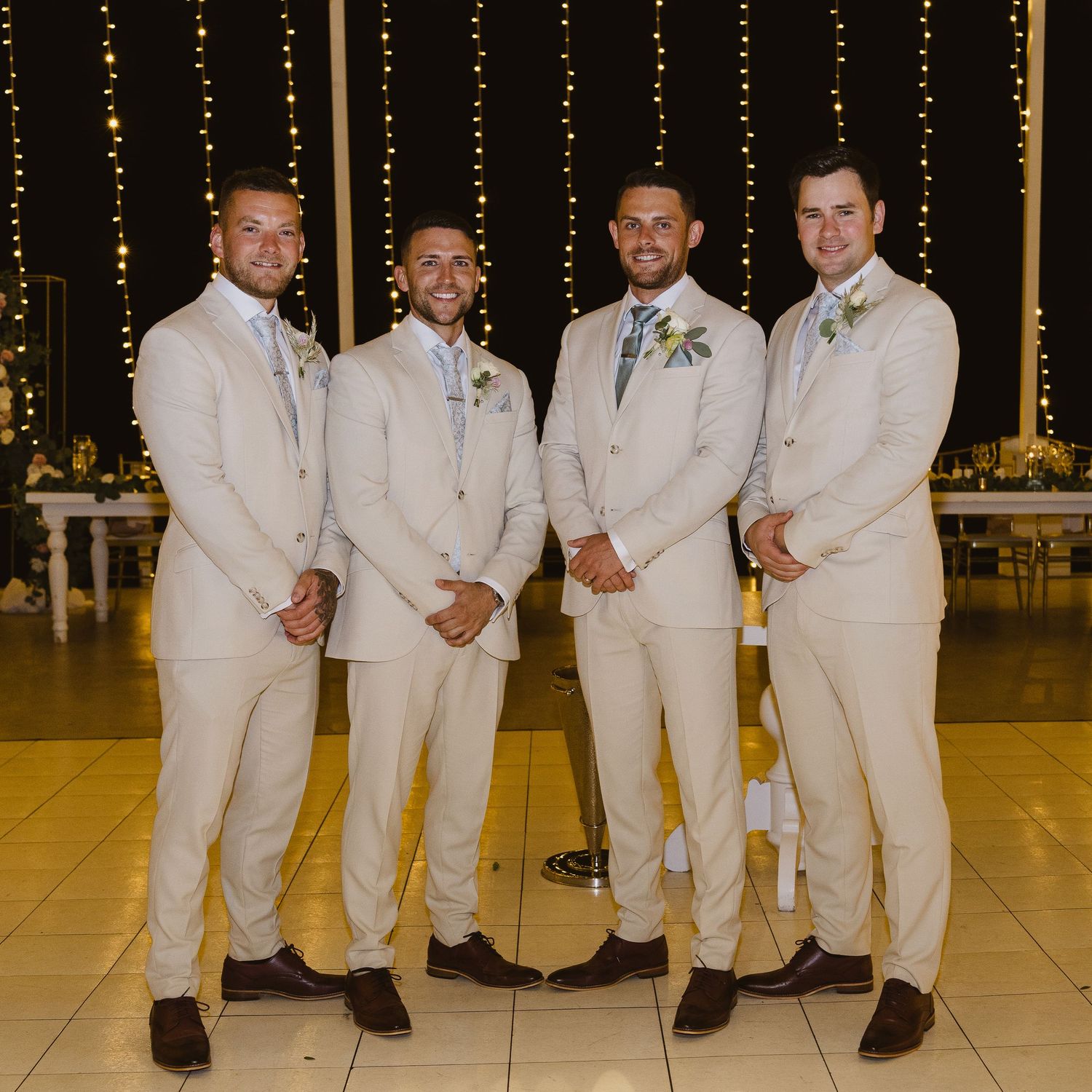 Groomsmen wearing light beige suits pose together at wedding reception.