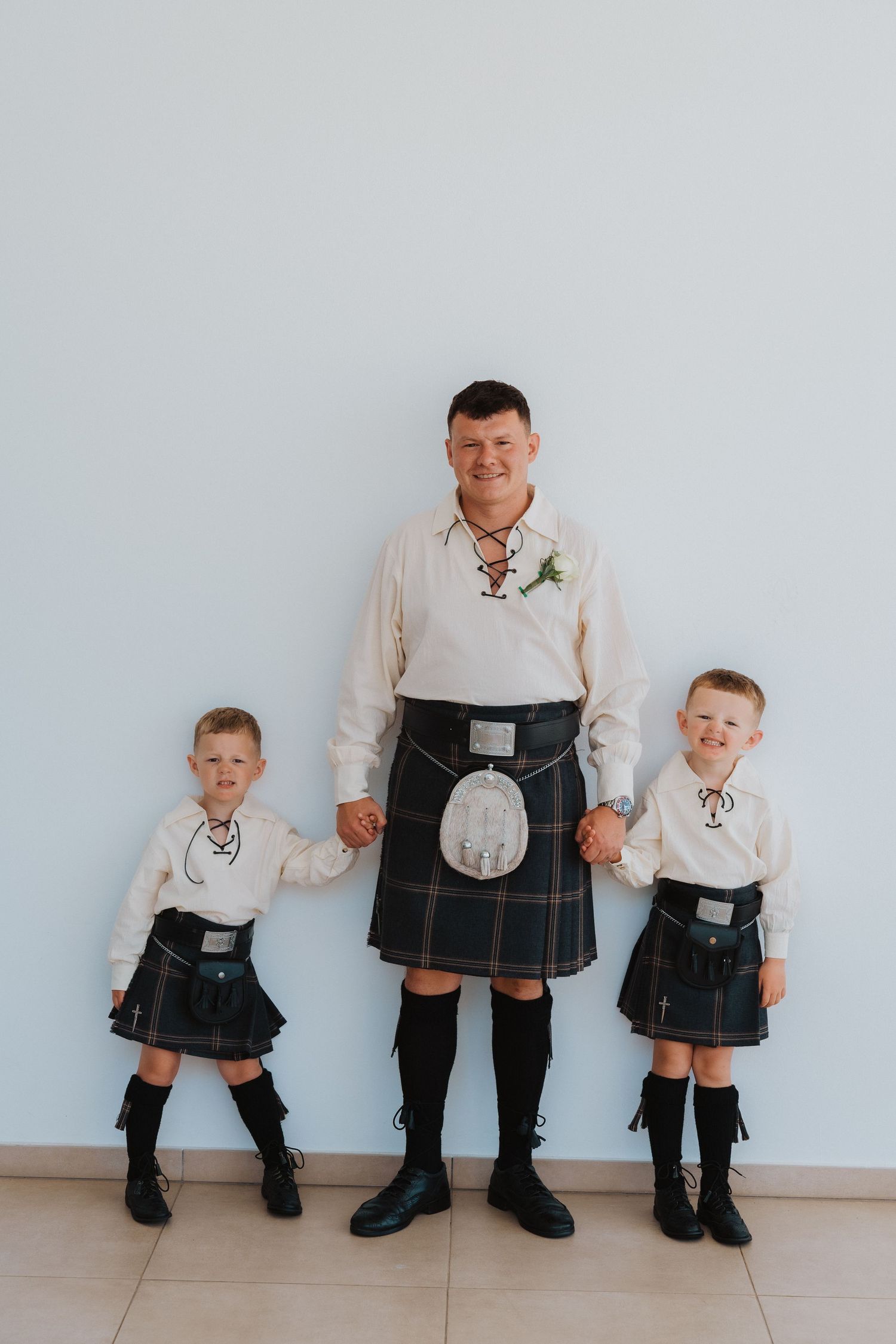 Three generations pose together in matching Scottish kilts and white dress shirts against a white wall.