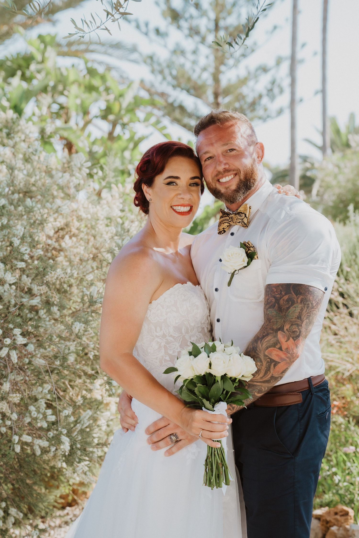 Bride holding white bouquet poses with groom in sunny garden setting.