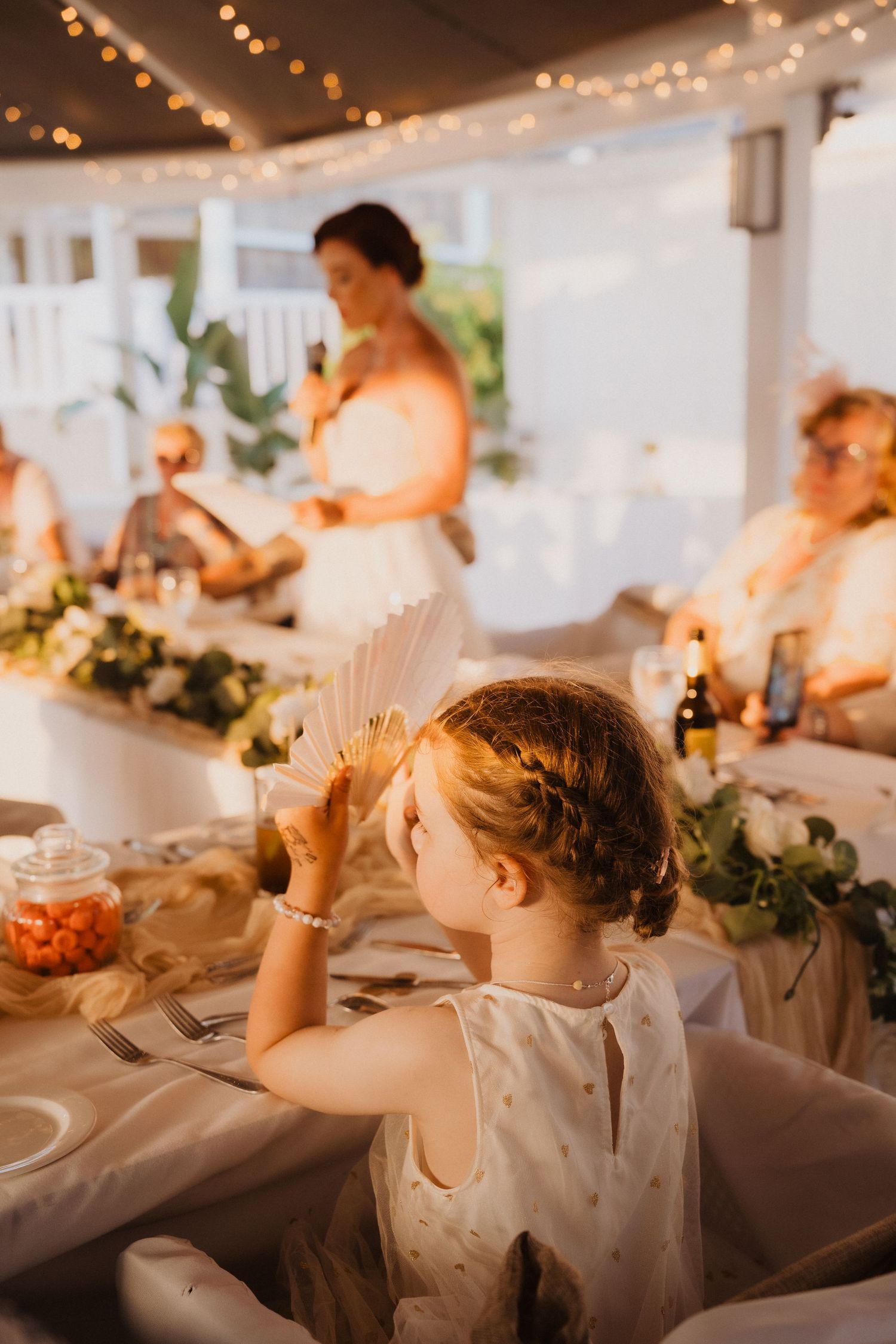 Child at wedding reception with string lights and floral decorations in background.