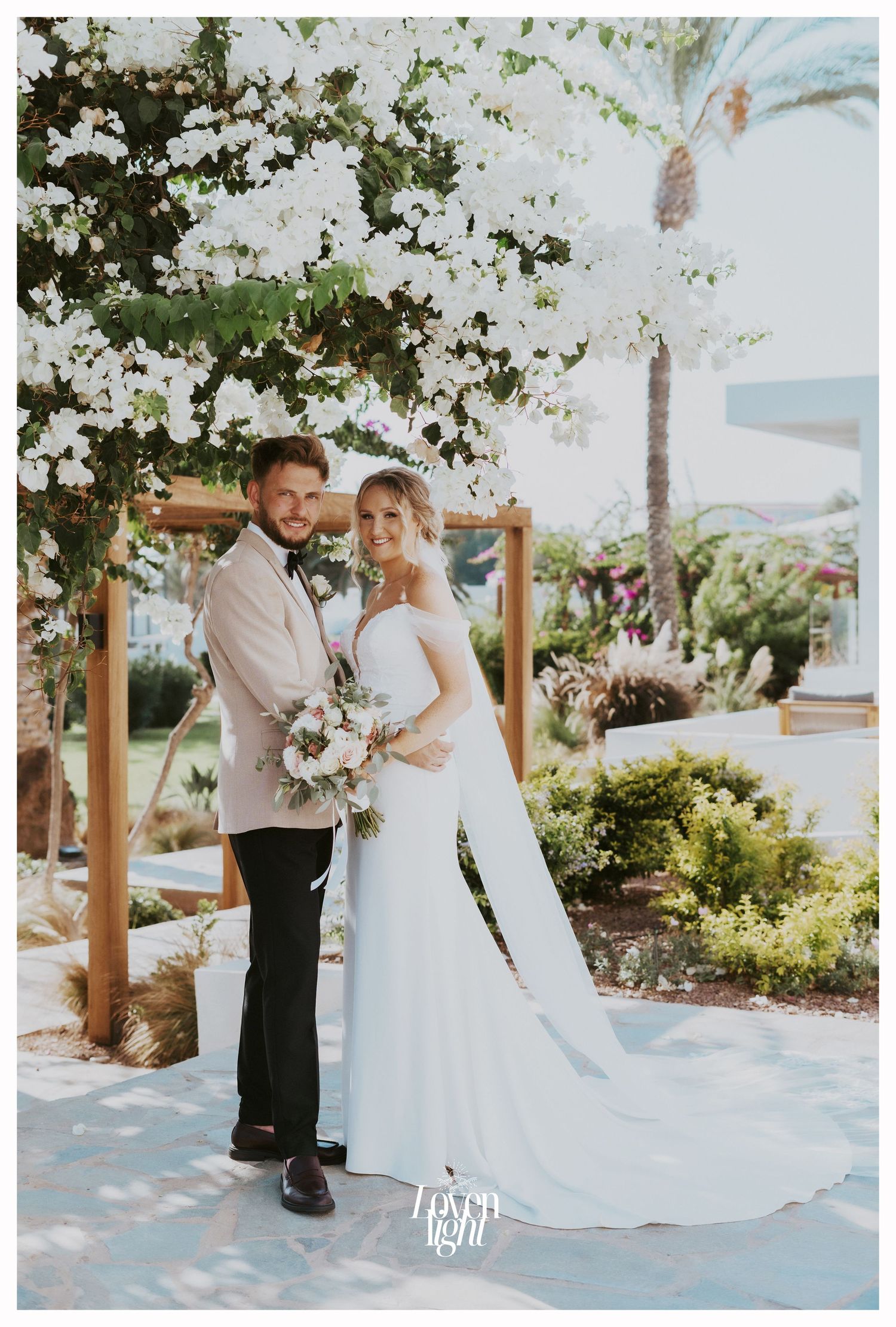 Wedding couple poses together under white flowering trees with palm trees in background.