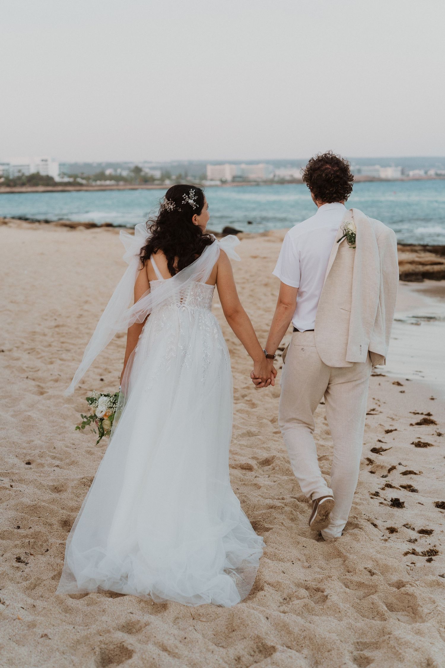 A wedding couple holding hands while walking on a sandy beach with turquoise waters in the background.