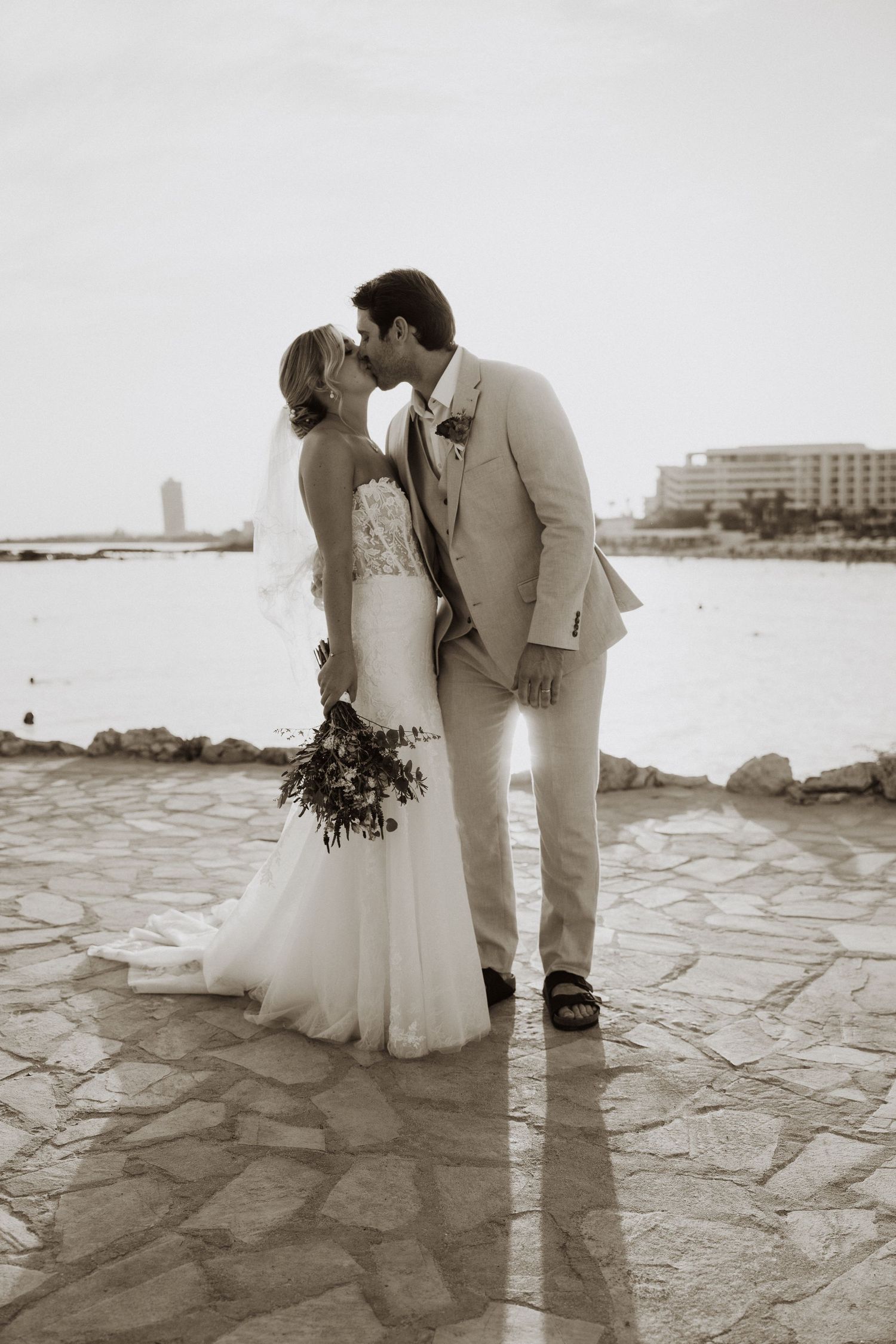 A black and white wedding photo of a couple kissing on a stone pathway by the water.