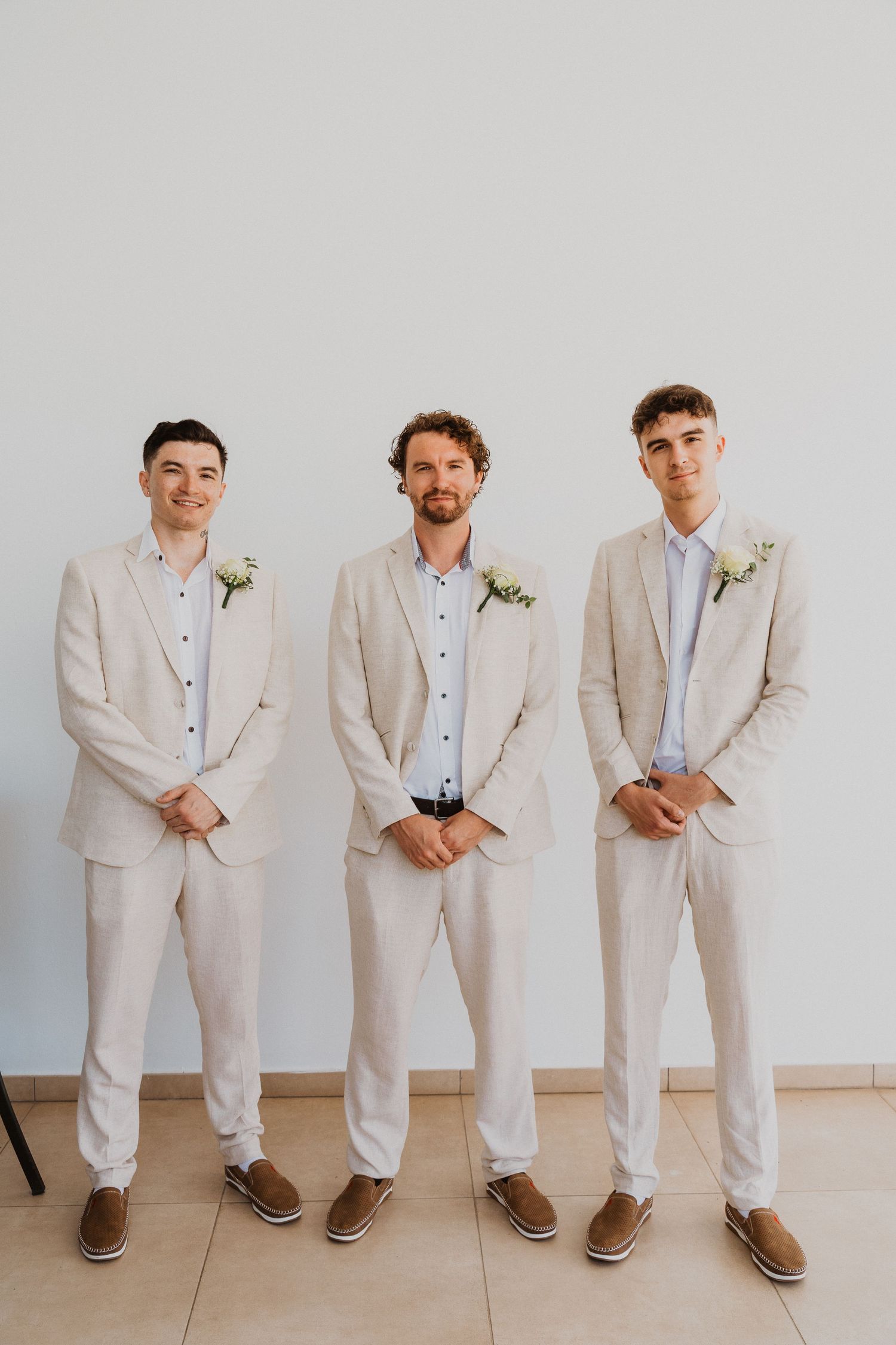 Three groomsmen in matching beige suits and boutonnieres stand against white wall.