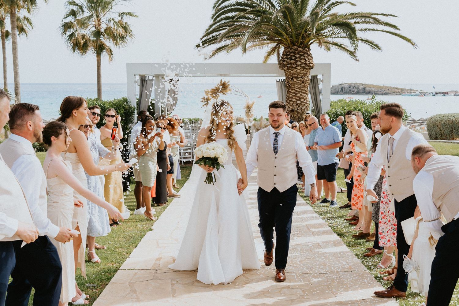 Wedding guests cheering along palm-lined path as newlyweds walk through.