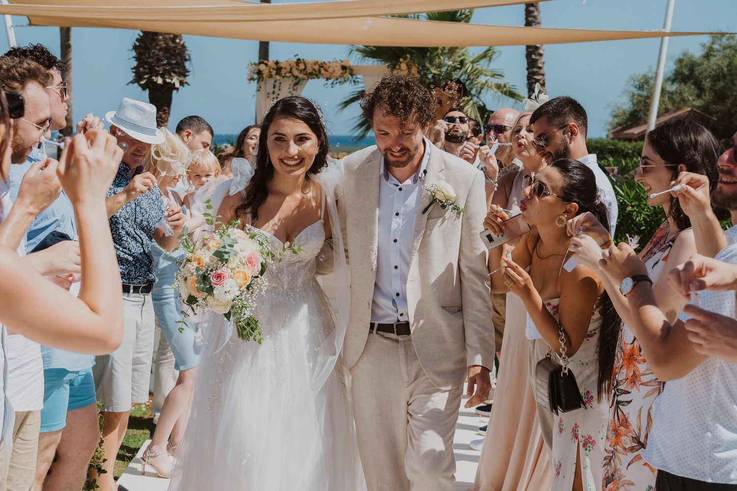 Beachside wedding celebration with guests throwing confetti as couple walks down outdoor aisle.