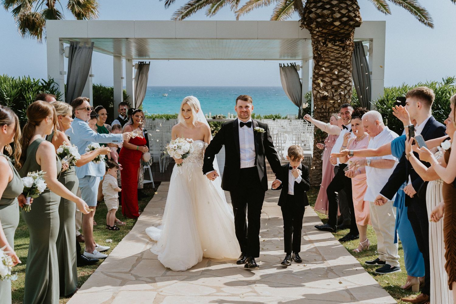 Guests applaud as newlyweds walk down an ocean-view aisle lined with palm trees.