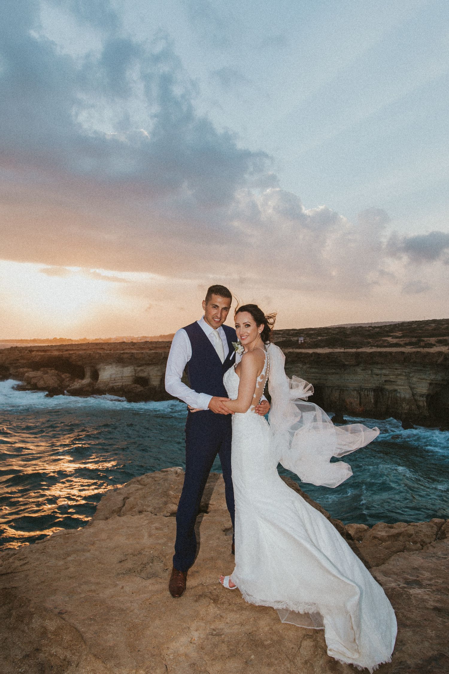 A dramatic sunset wedding photo with cliffs and ocean backdrop as dress flows in wind.