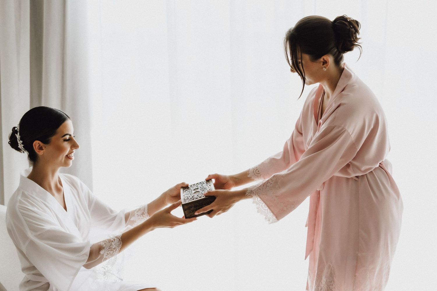 Person in pink robe hands something to person wearing white robe in bright room setting.