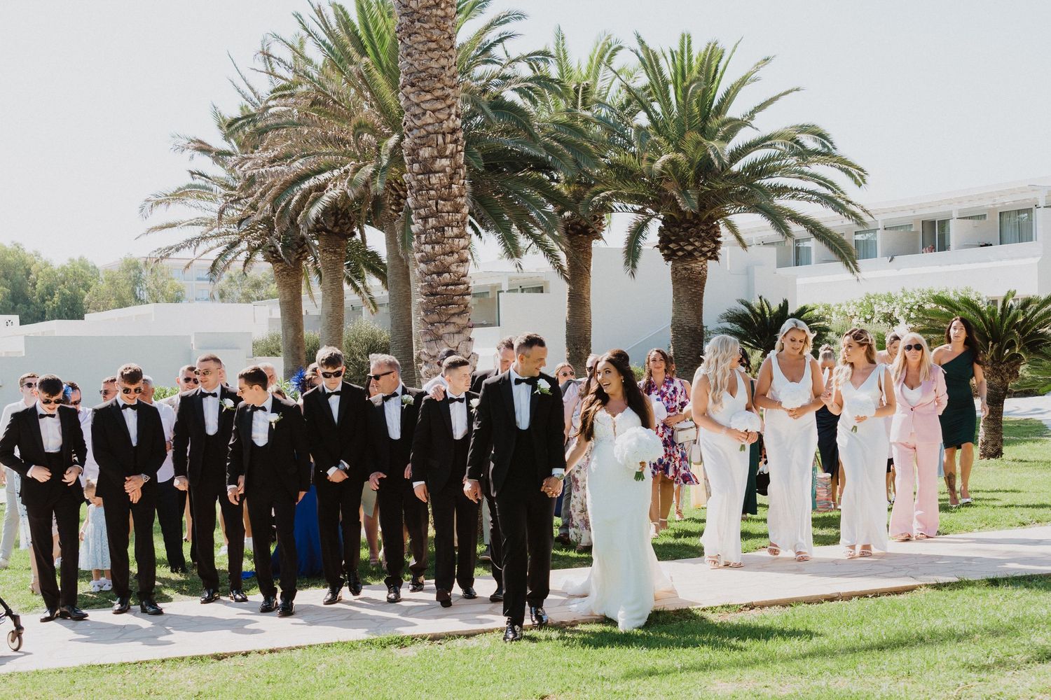Wedding party members arranged in formal groups on a sunny lawn with palm trees.