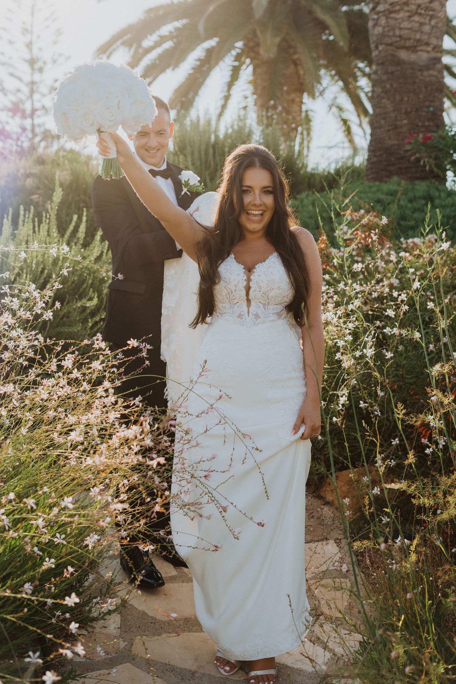 A bride in a white dress twirls joyfully among palm trees and wildflowers in a garden setting.