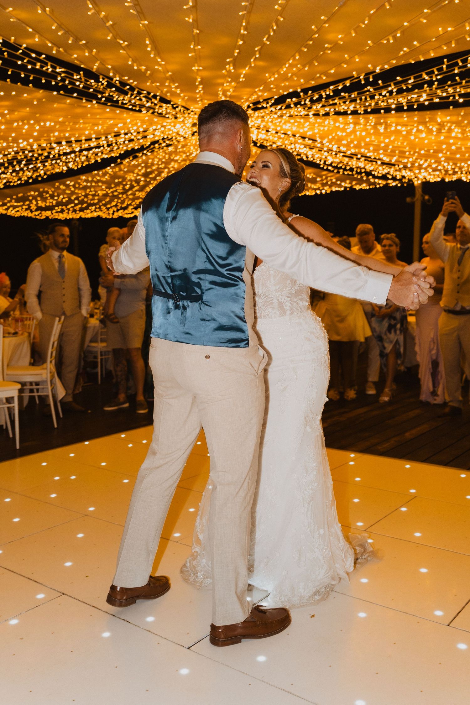 A couple shares their first dance under a canopy of twinkling golden lights at an outdoor evening wedding reception.