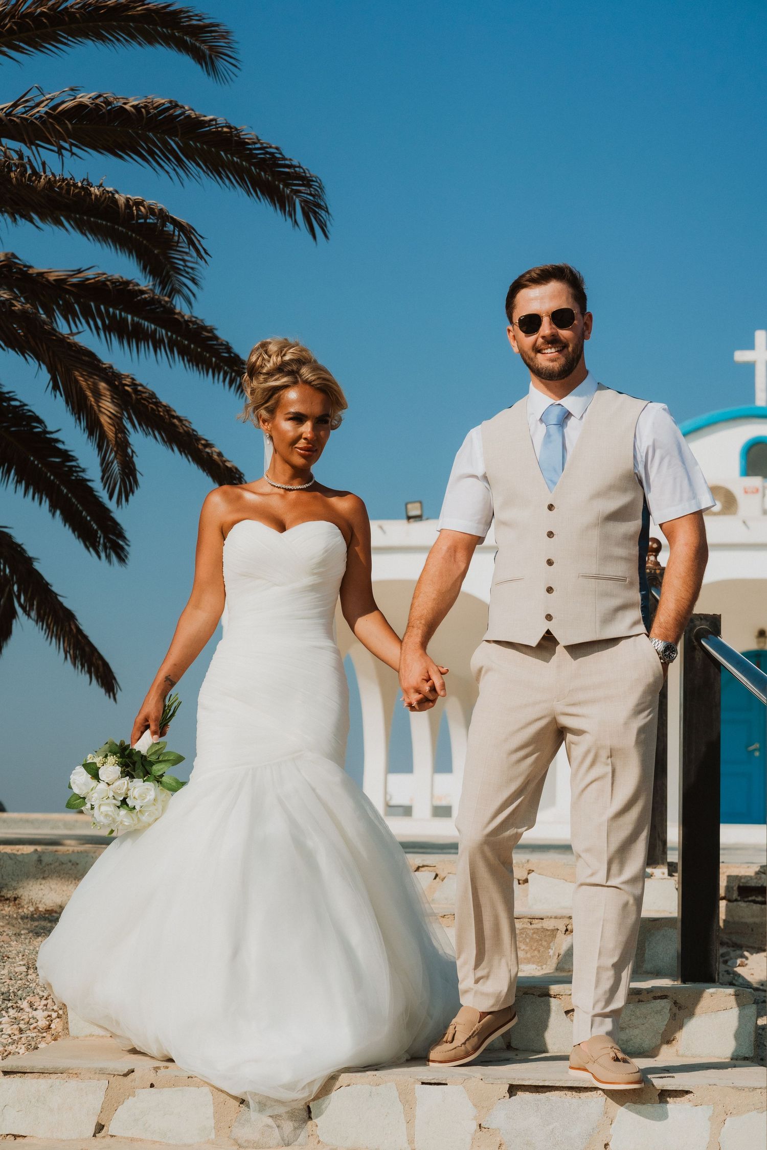 Bride and groom holding hands outdoors in Santorini, white church and palm tree in background on sunny day.