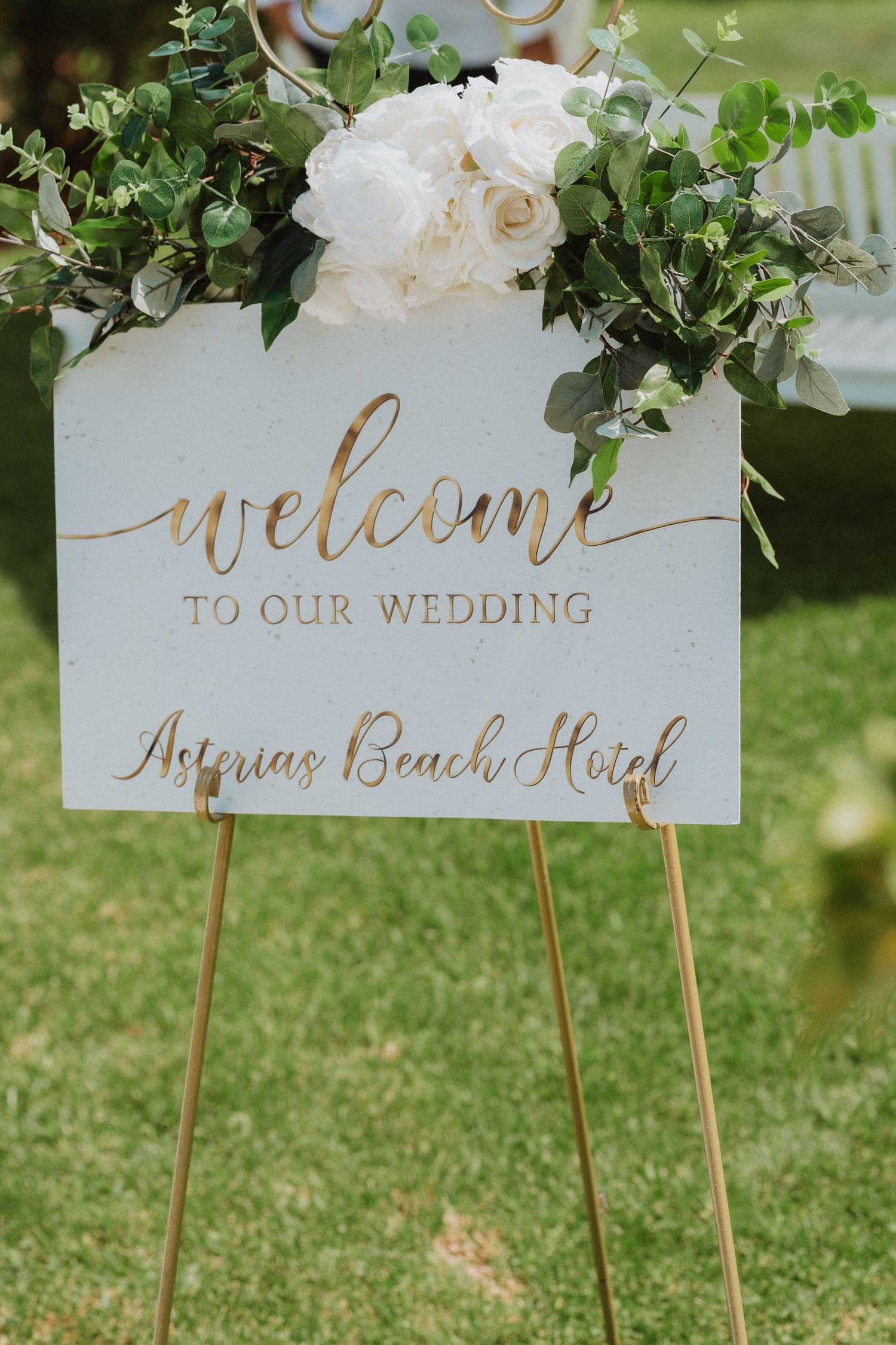Elegant wedding welcome sign at Asterias Beach Hotel with gold script lettering and white floral arrangement on gold easel.