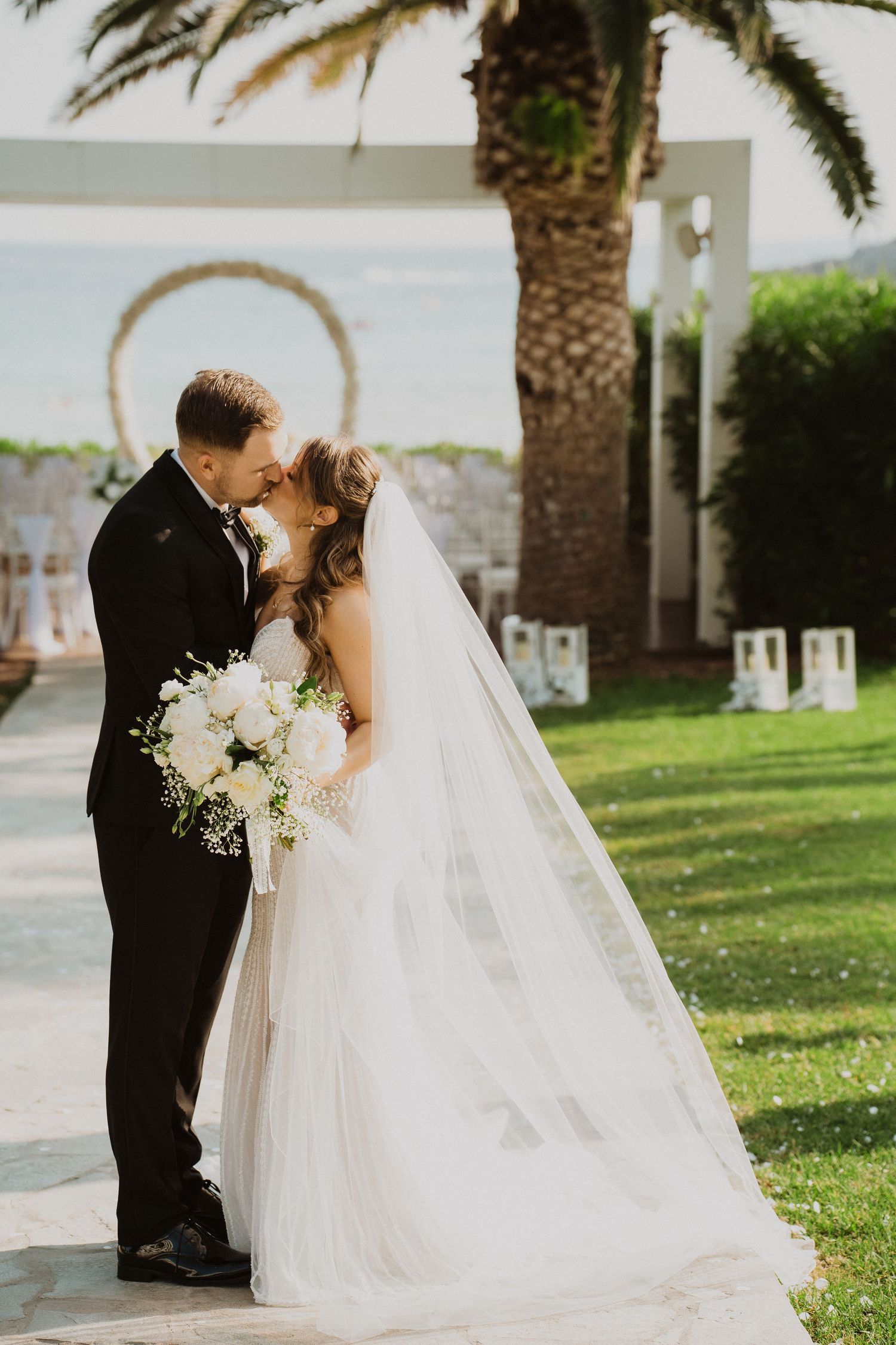 Bride and groom sharing romantic moment on garden path, bride holding white bouquet with long veil flowing.
